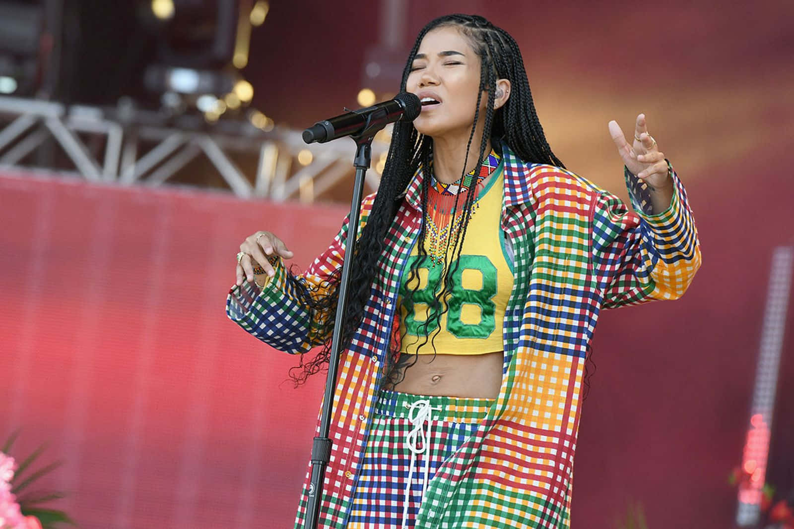 jhene aiko 桌布 壁紙 — 一位充滿活力的表演者在明亮的舞台燈光下，穿著色彩繽紛的格子服裝，熱情地唱歌。