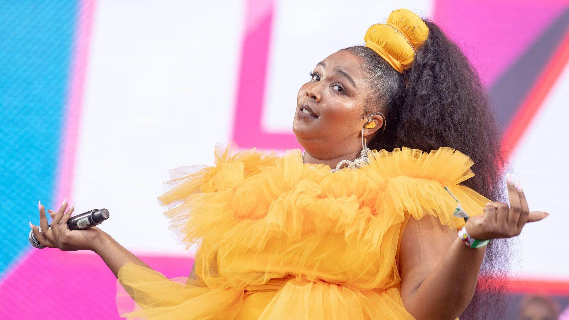 Lizzo 桌布 壁紙 — 一位充滿活力的表演者身穿荷葉邊黃色連衣裙，在色彩繽紛的舞台上熱情歌唱，身後是醒目的粉紅色和藍色幾何燈光。