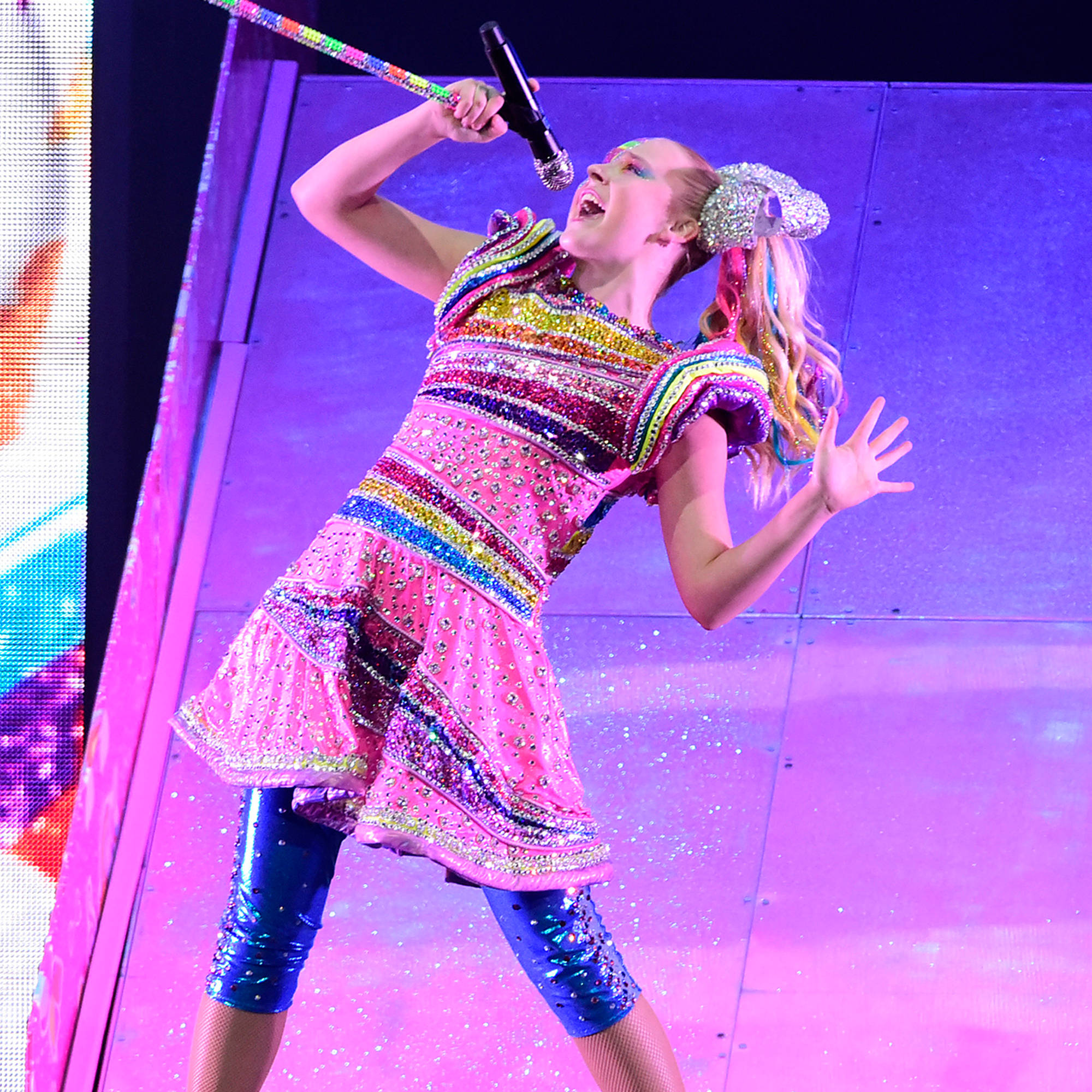 JoJo Siwa 桌布 壁紙 — 一位充滿活力的表演者身穿閃閃發光的彩虹服裝，在紫色燈光的舞台上激情演唱，火花四濺。