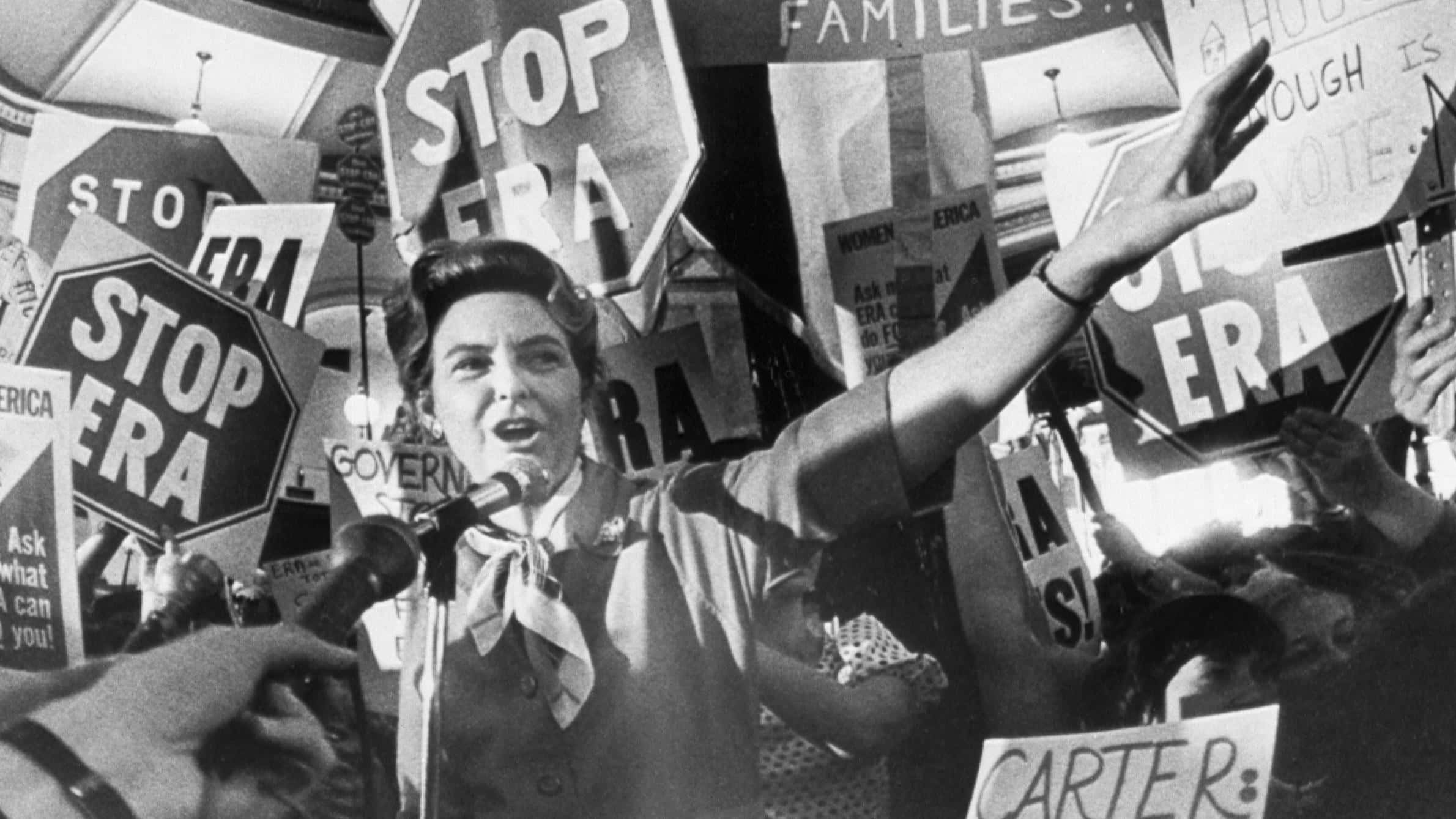 Politics 桌布 壁紙 — 一位充滿熱情的女性對著麥克風說話，周圍是舉著「STOP ERA」標語的抗議者，在擁擠的集會中。