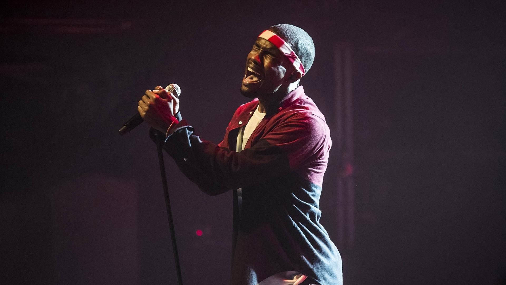 Frank Ocean 桌布 壁紙 — 一位充滿熱情的男歌手在帶有紅色和紫色色調的戲劇性舞台燈光下對著麥克風高歌。