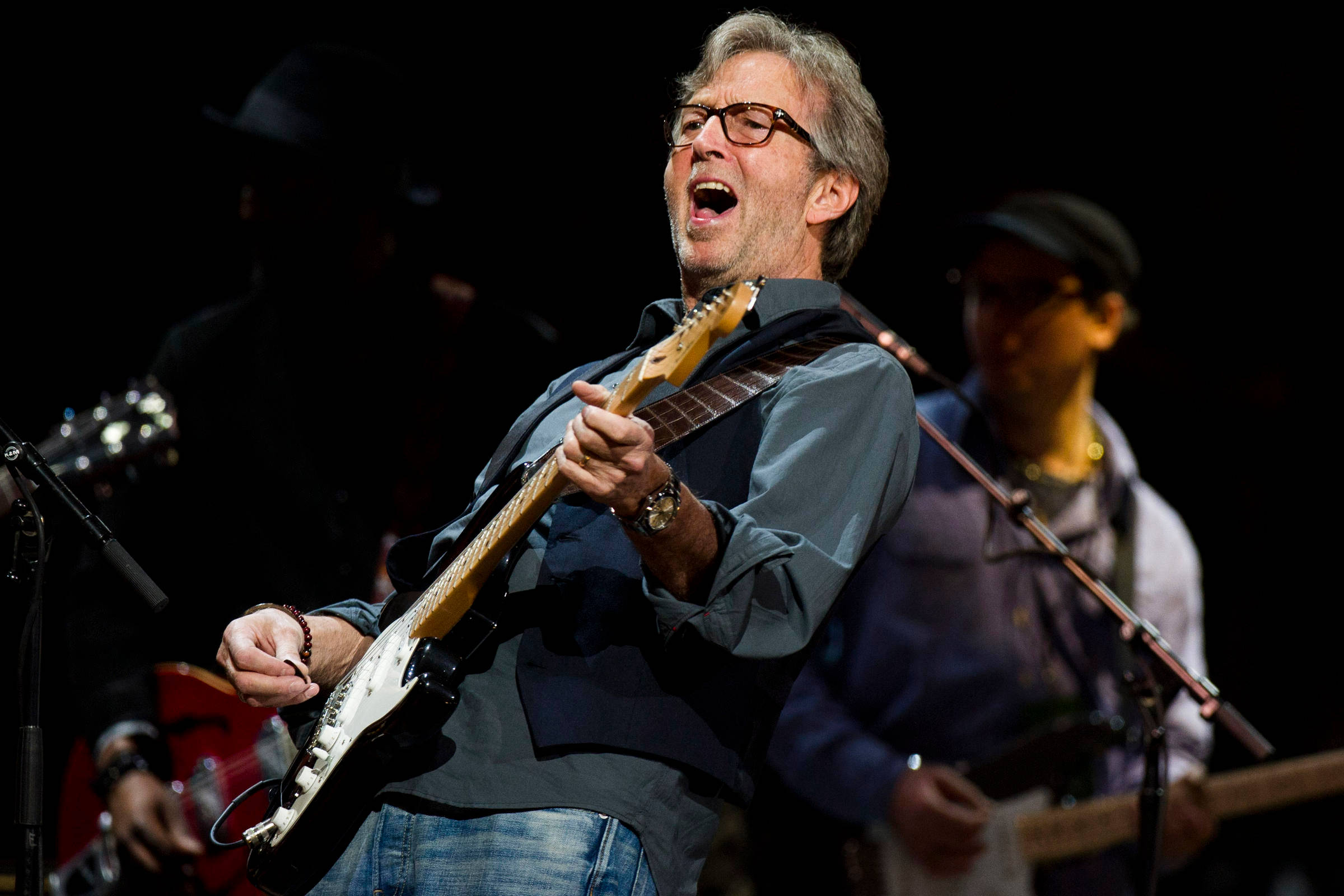 guitar player 桌布 壁紙 — 一位充滿熱情的音樂家戴著眼鏡，在明亮的聚光燈下熱情地彈奏電吉他，背景中的樂隊成員模糊不清。