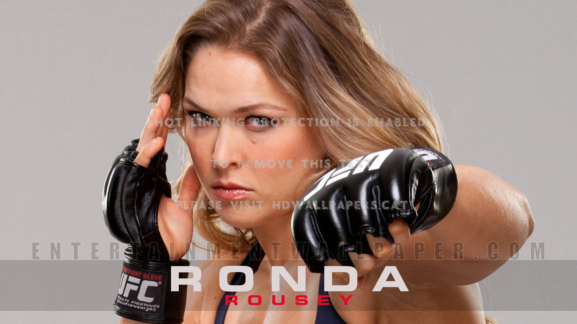 Ronda Rousey 桌布 壁紙 — 一位兇猛的女戰士戴著黑色UFC手套，在純灰色背景前舉起一隻手擺出防禦姿勢，眼神銳利。