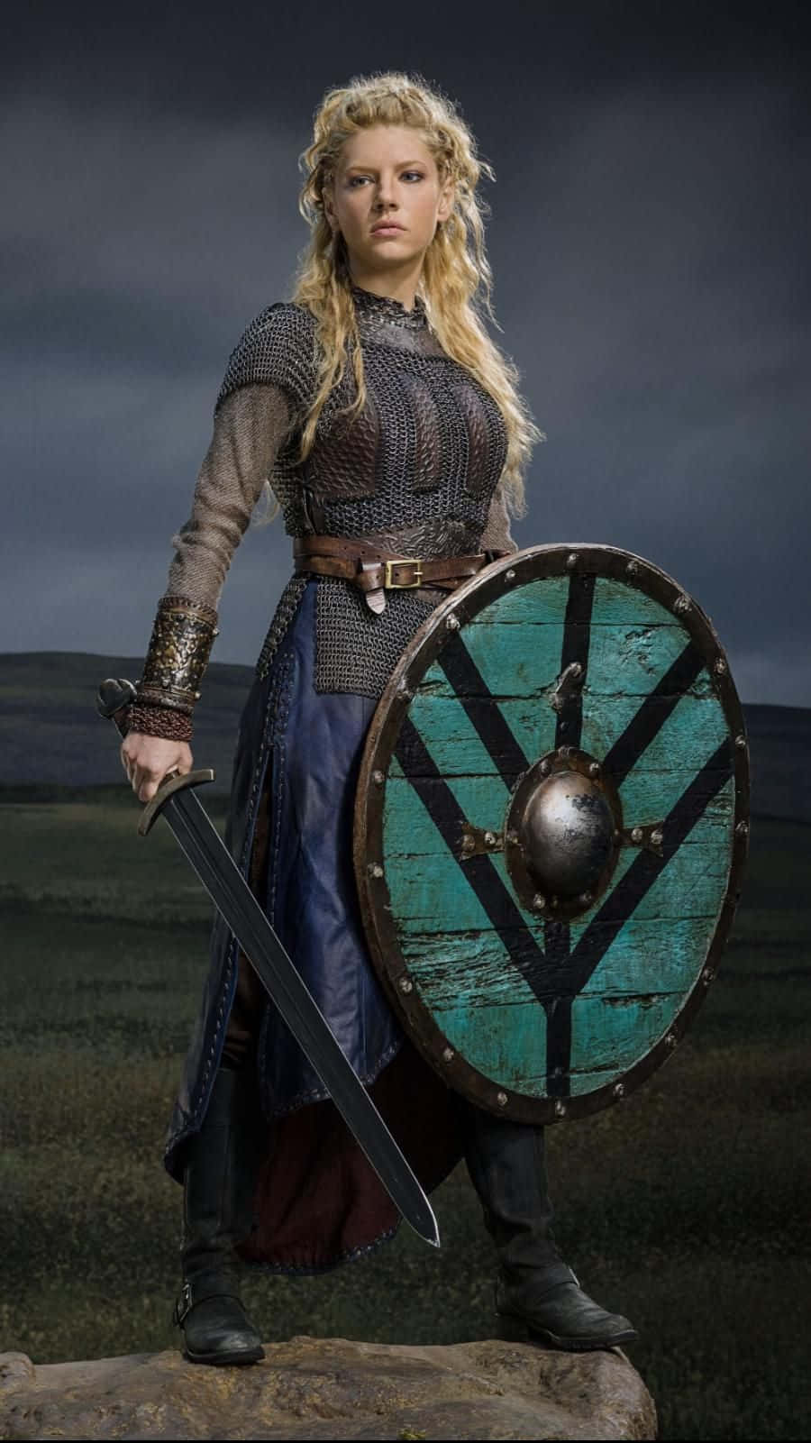 Female Viking Warriors 桌布 壁紙 — 一位兇猛的金髮維京女子，身穿鎖子甲和藍色裙子，站在岩石上，手持劍和青綠色盾牌，背景是風暴天空。