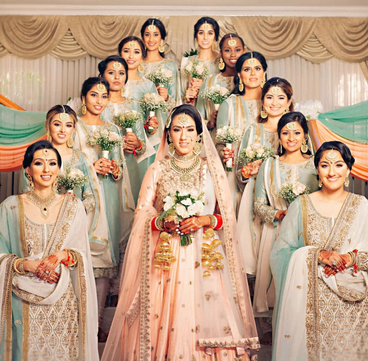 Bridesmaids 桌布 壁紙 — 一位光彩奪目的新娘身穿華麗的粉紅色和金色 lehenga，站在舞台中央，周圍是她的伴娘，她們穿著相配的淺藍色和金色傳統服飾，手持白色花束。