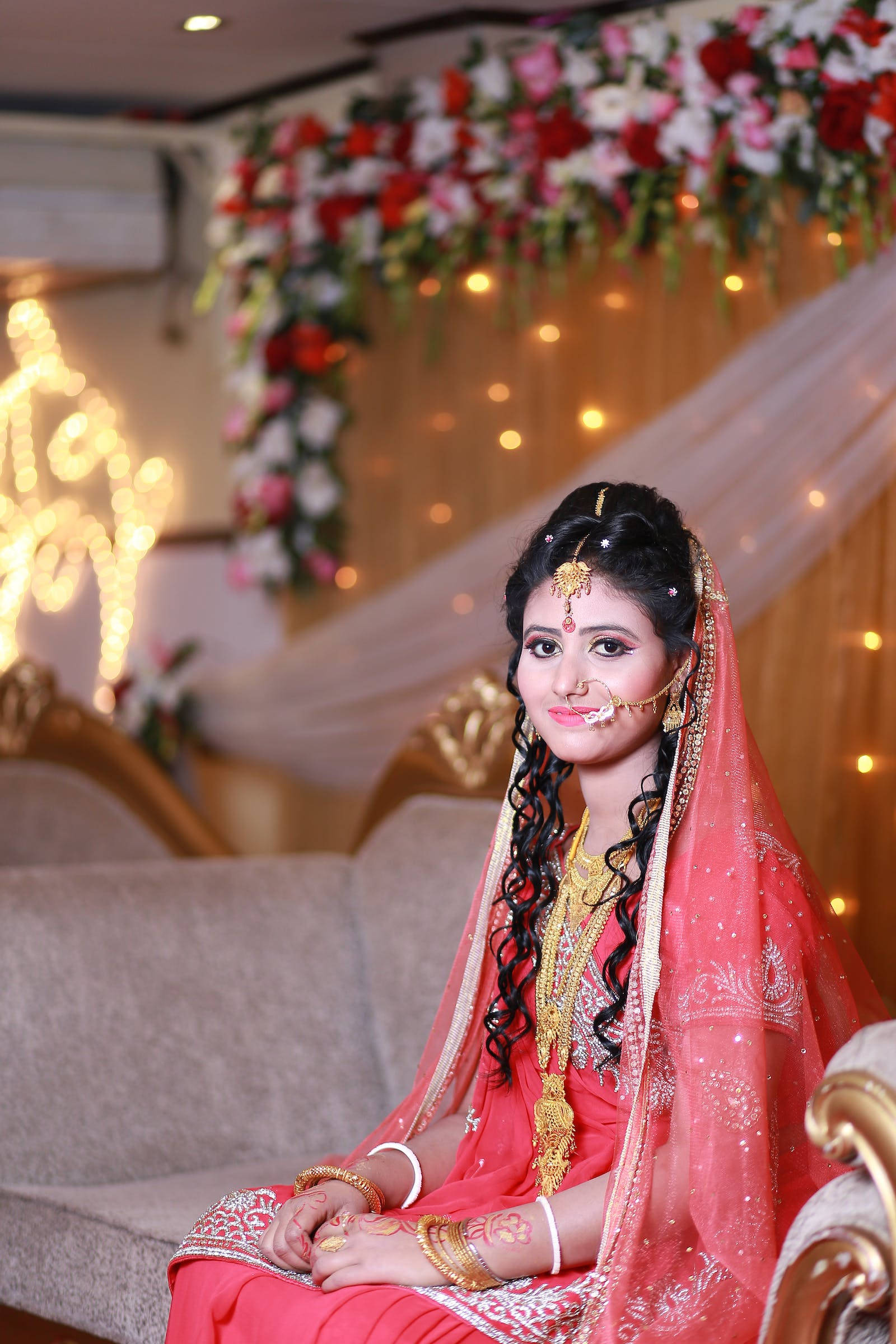 Indian 桌布 壁紙 — 一位光彩奪目的新娘身穿鮮豔的粉紅色 lehenga，佩戴金色珠寶，優雅地坐在花卉拱門和閃爍的仙女燈下。
