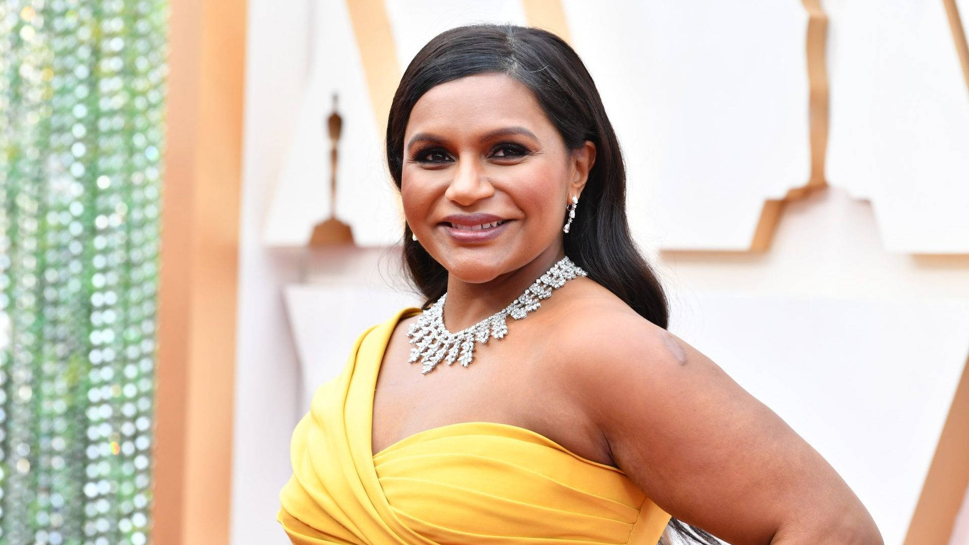 oscars 桌布 壁紙 — 一位光彩照人的女士身穿鮮黃色禮服，佩戴閃爍的鑽石項鍊，在華麗的紅地毯上自信地微笑。