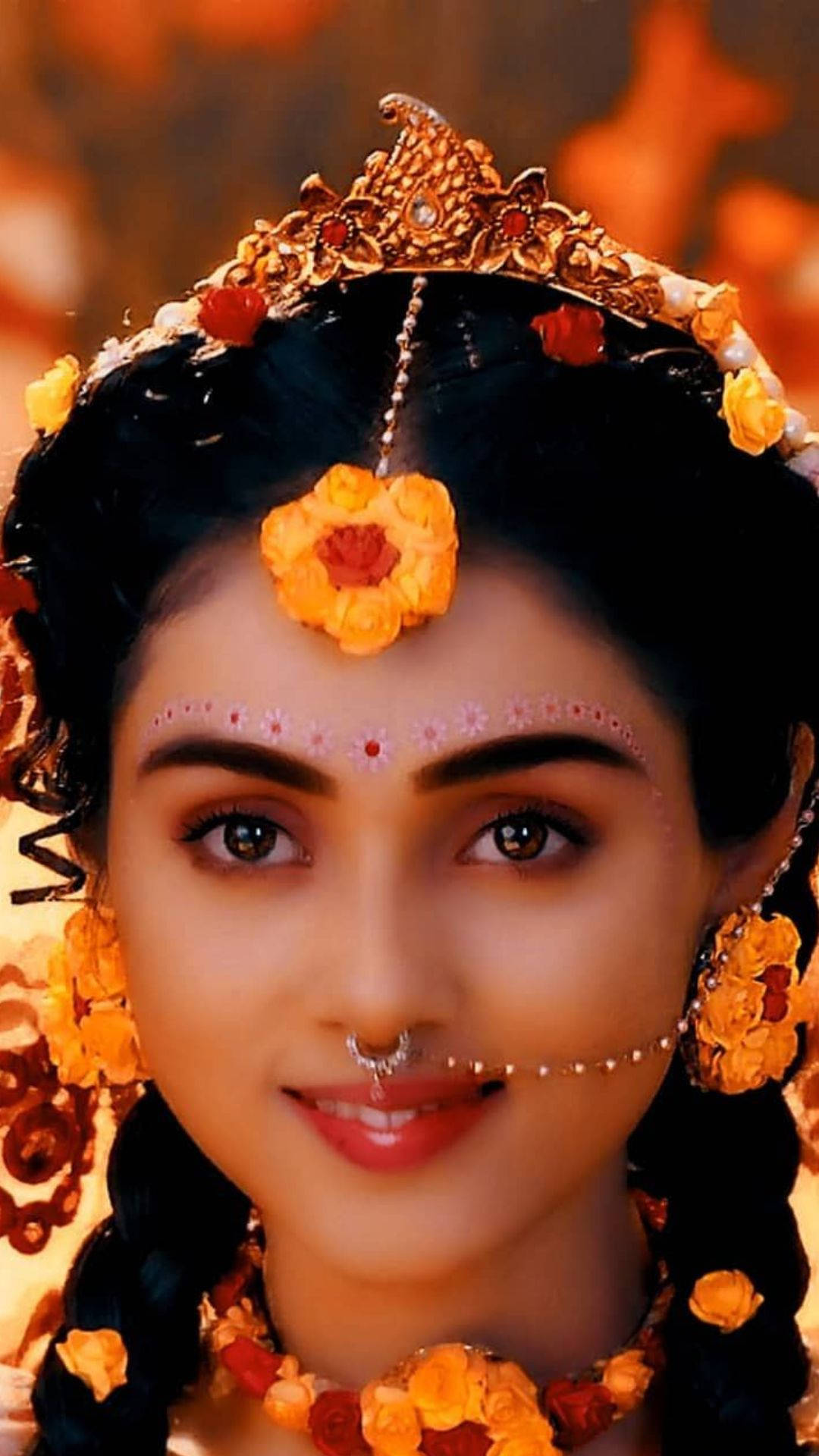 radha krishna serial 桌布 壁紙 — 一位光彩照人的女性，佩戴金色花冠和珠寶，在發光的橙色背景下溫暖地微笑。