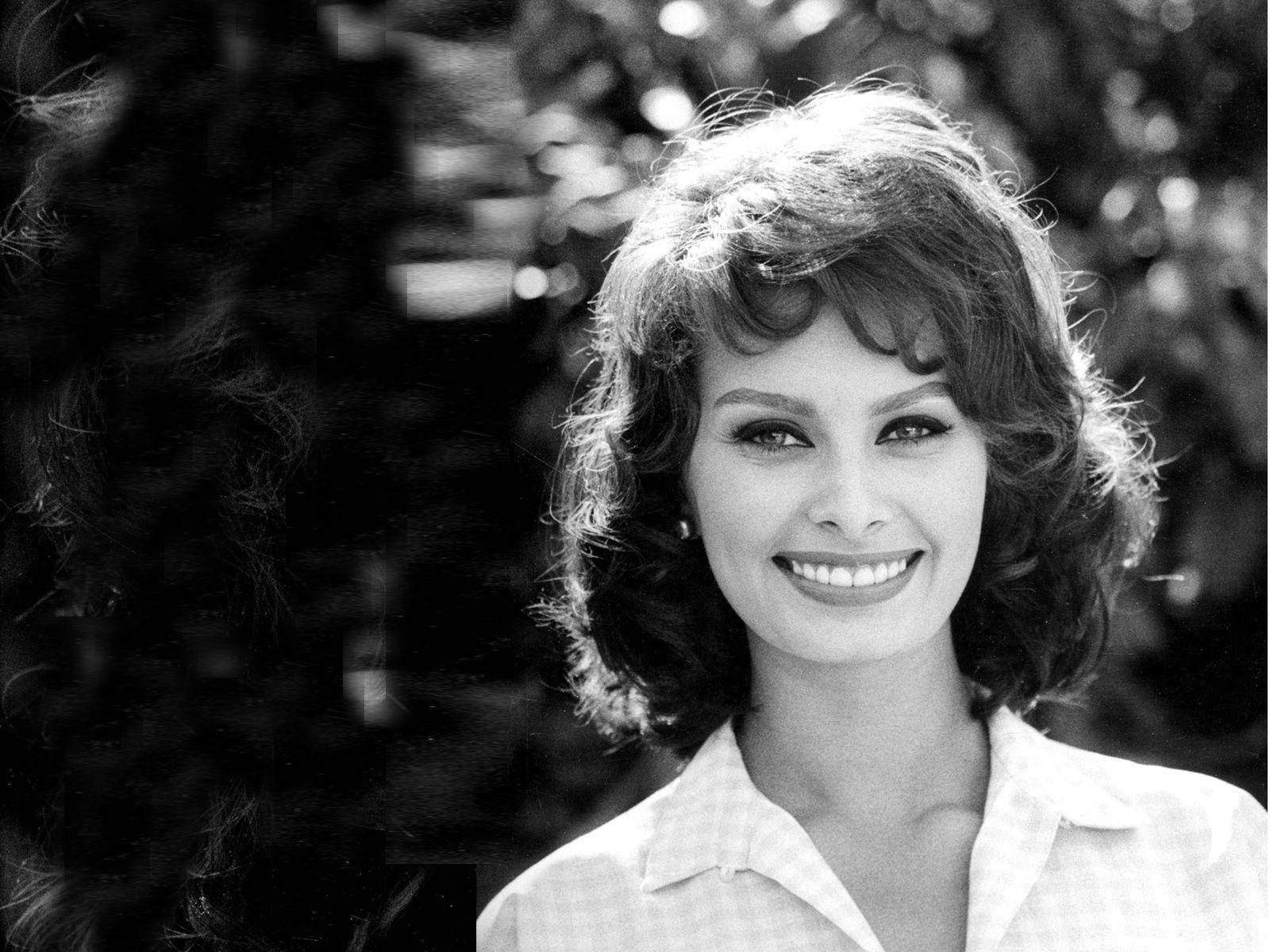 Actress HD 桌布 壁紙 — 一位光彩照人的女性，擁有豐盈的捲髮，在經典黑白背景下，對著柔和模糊的樹葉背景溫暖微笑。