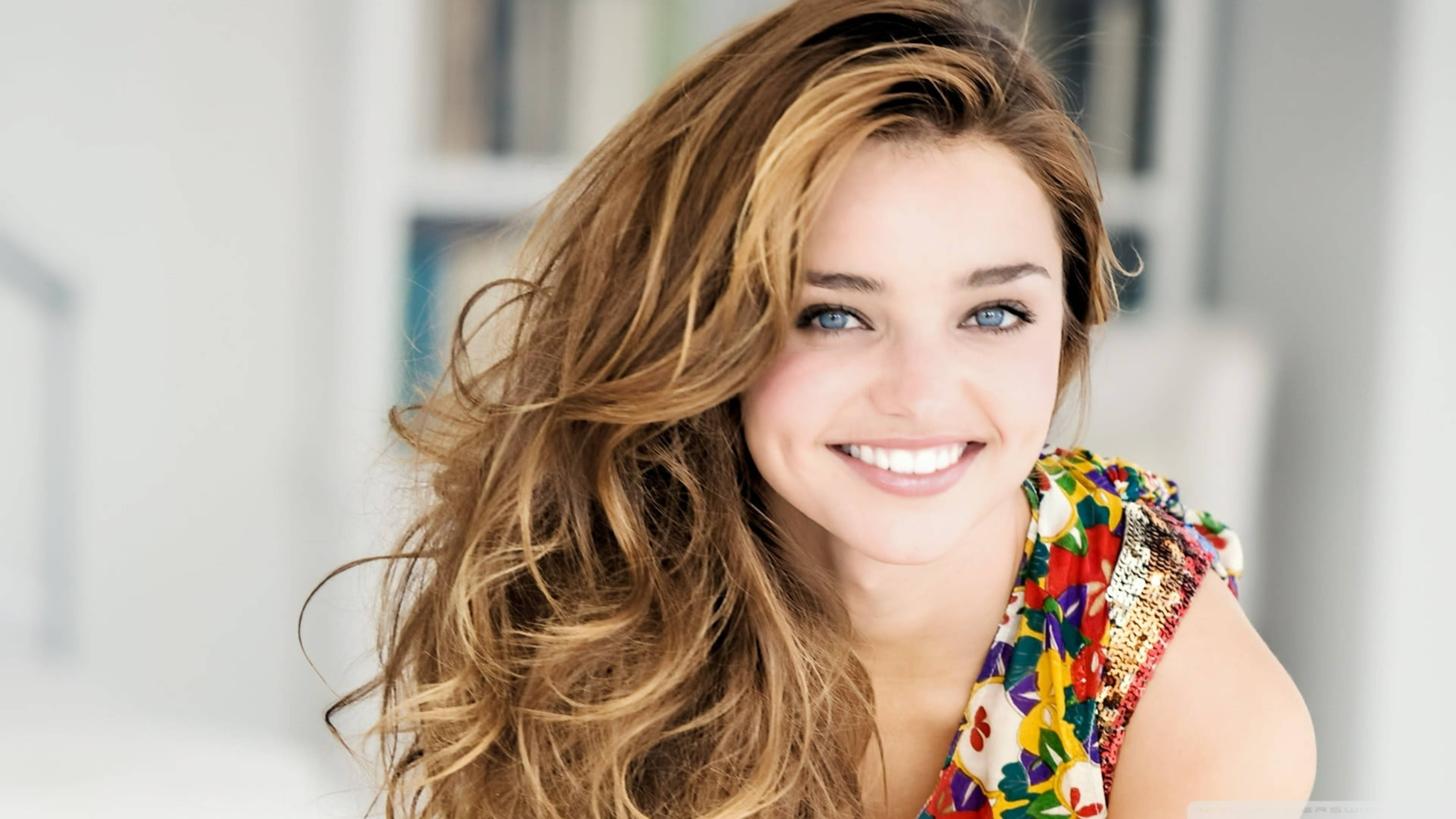 Miranda Kerr 桌布 壁紙 — 一位光彩照人的女性，擁有飄逸的金色長髮和明亮的藍眼睛，在陽光充足的房間裡穿著色彩繽紛的花卉上衣，溫暖地微笑。