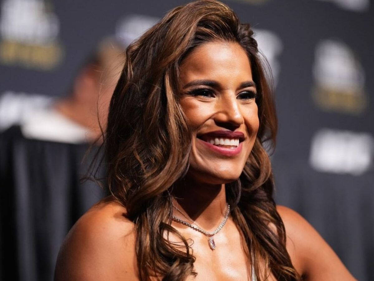 UFC 桌布 壁紙 — 一位光彩照人的女性，波浪棕色頭髮，在深色品牌背景的活動中燦爛微笑。
