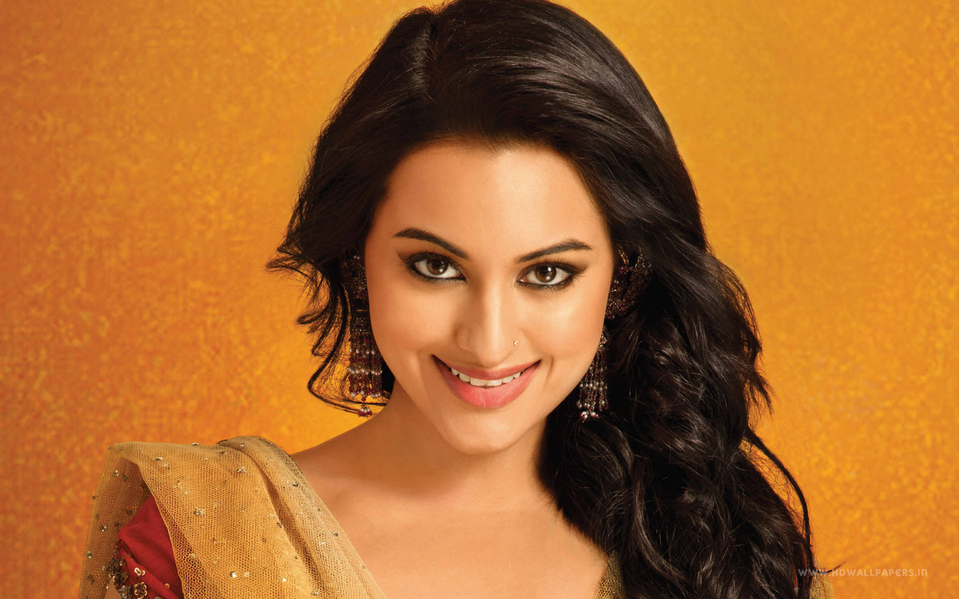 Sonakshi Sinha 桌布 壁紙 — 一位光彩照人的女性，深色波浪長髮，佩戴傳統珠寶，在帶有紋理的金色背景前溫暖微笑。