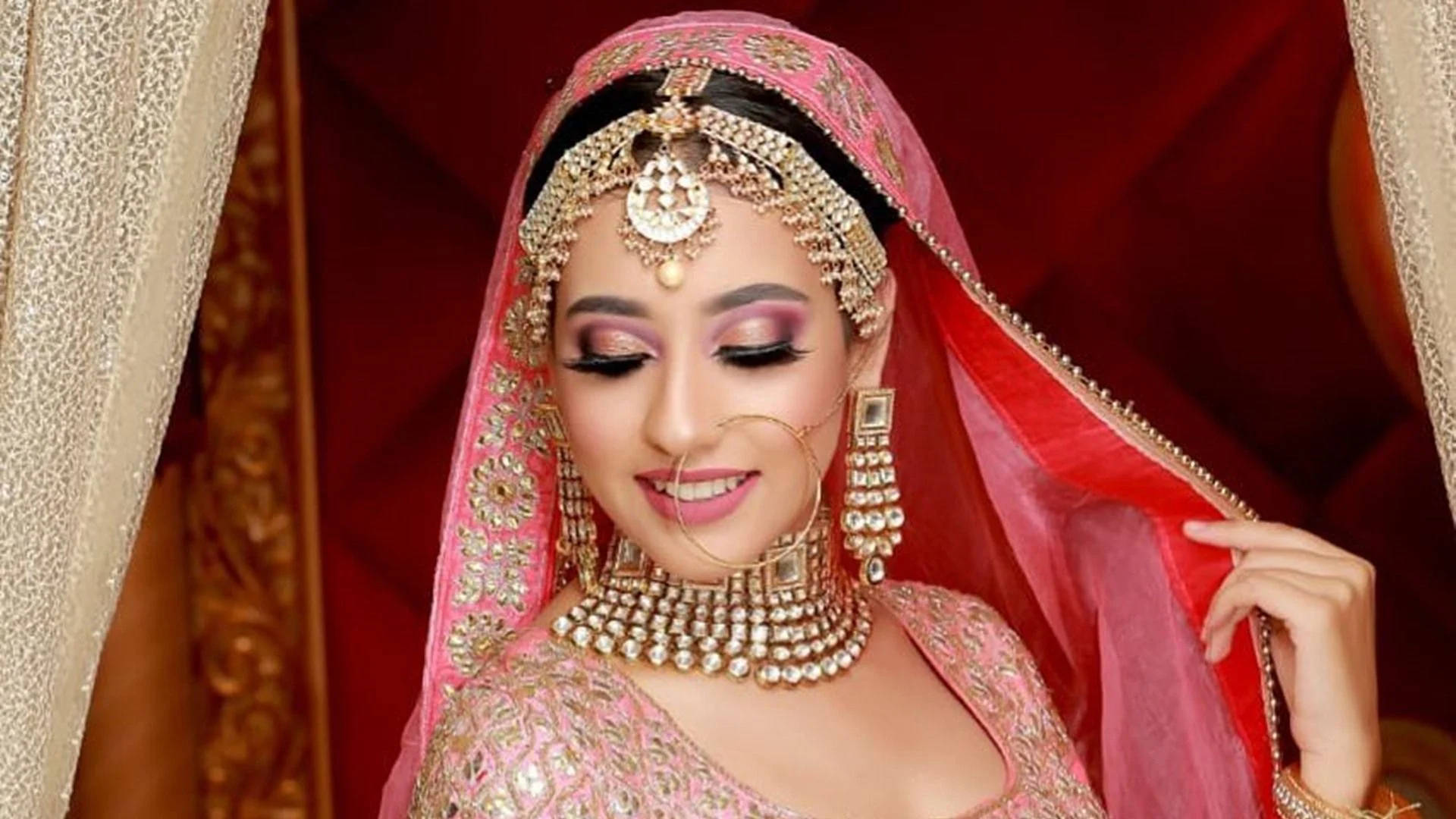 Bridal 桌布 壁紙 — 一位光彩照人的新娘，身穿華麗的粉紅色 lehenga 和黃金首飾，在濃郁的紅色背景下溫柔微笑。