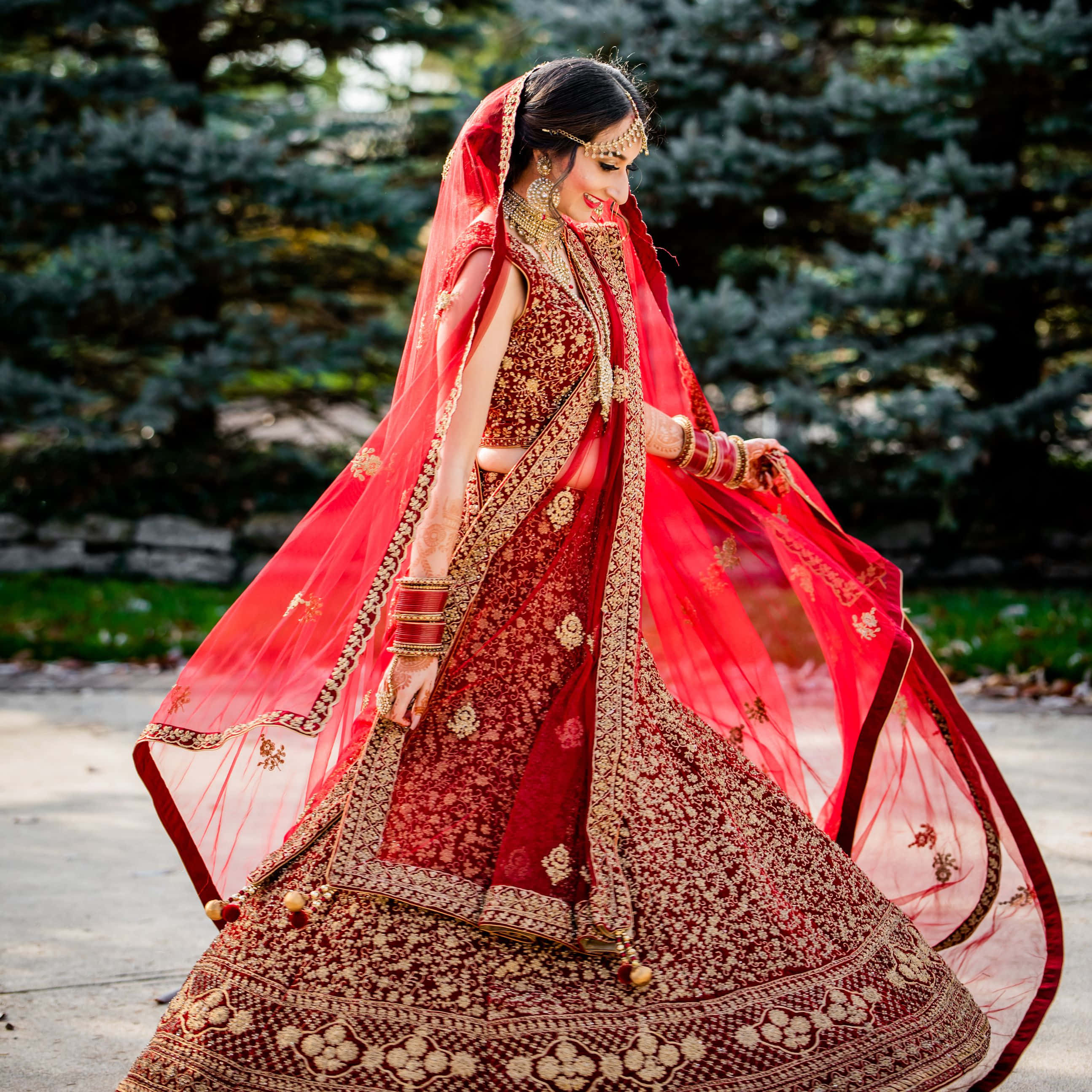 Bride 桌布 壁紙 — 一位光彩照人的新娘穿著華麗的紅色和金色刺繡 lehenga，披著飄逸的面紗，優雅地在戶外行走。