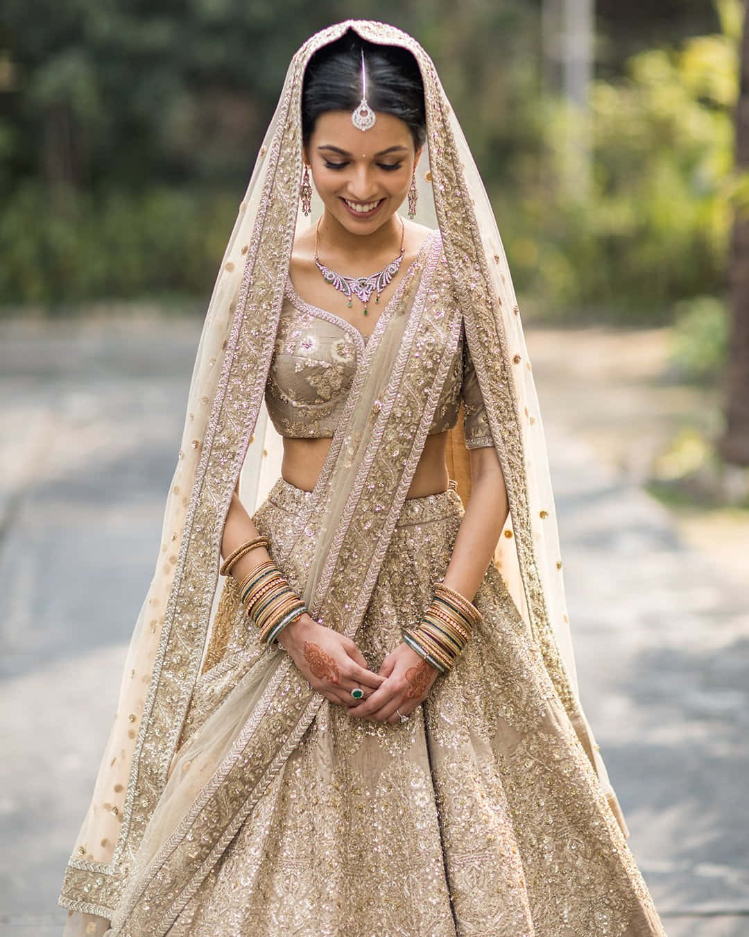 Smile 桌布 壁紙 — 一位光彩照人的新娘穿著華麗的金色 lehenga，帶有精緻刺繡和透明面紗，在戶外微笑。