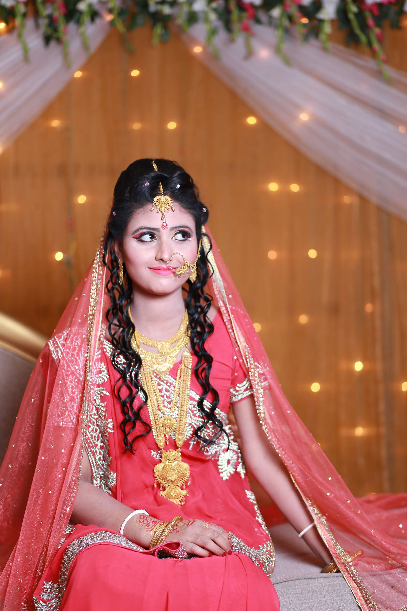 Indian 桌布 壁紙 — 一位光彩照人的新娘身穿鮮豔的粉紅色 lehenga 和金色珠寶，在掛有閃爍燈光的垂簾天篷下溫柔微笑。