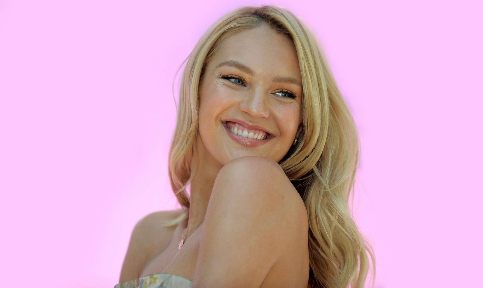 Candice 桌布 壁紙 — 一位光彩照人的金髮女郎在純粉紅色背景和柔和燈光下溫暖地微笑。
