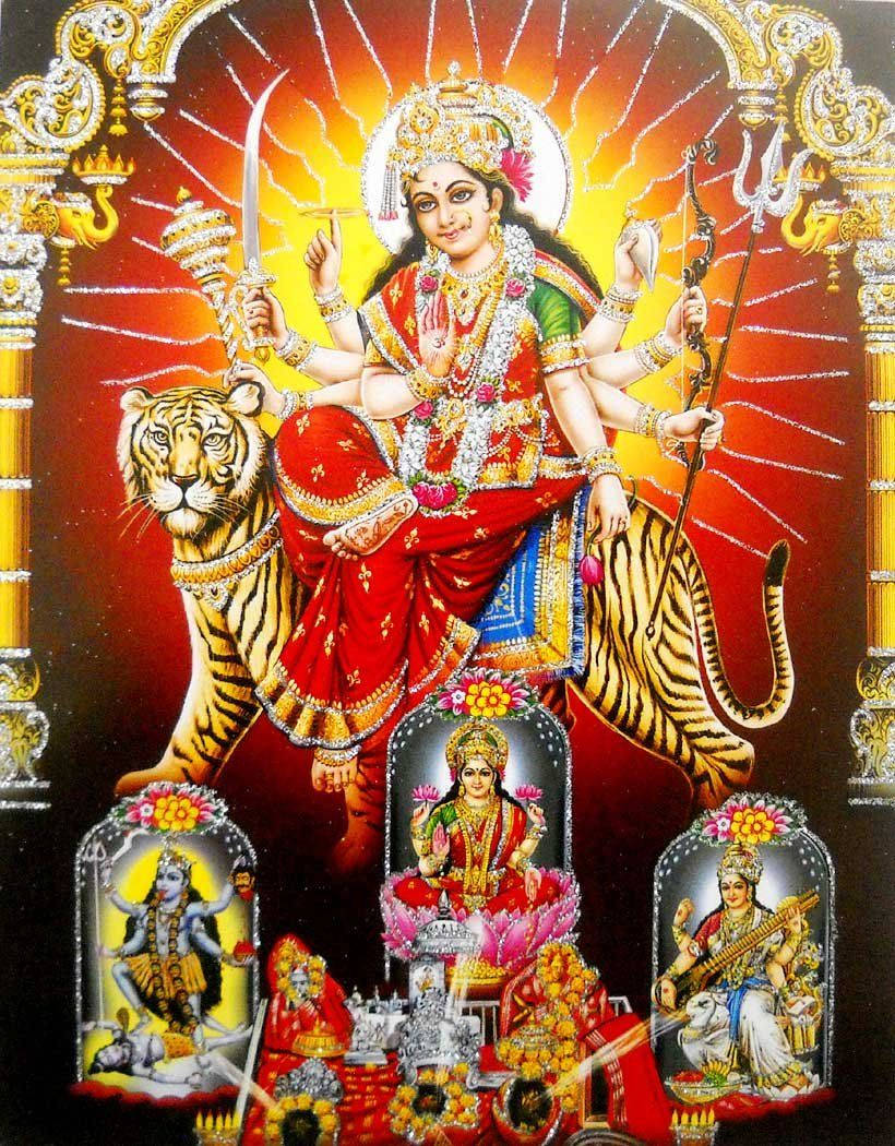 Vaishno Devi 桌布 壁紙 — 一位光芒四射、多臂的杜爾迦女神身穿紅金色，坐在老虎上，手持武器，下方壁龕中有神聖人物。