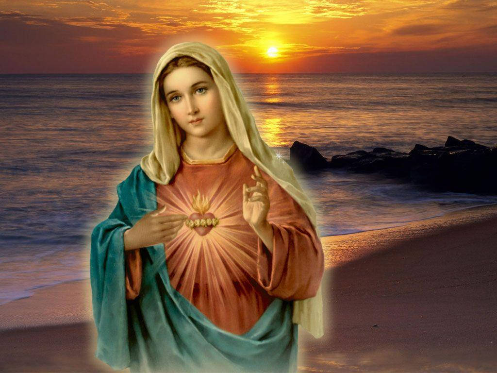Virgin Mary 桌布 壁紙 — 一位光芒四射的宗教人物，帶著發光的聖心，站在日落時分的海灘上，海浪在附近拍打。