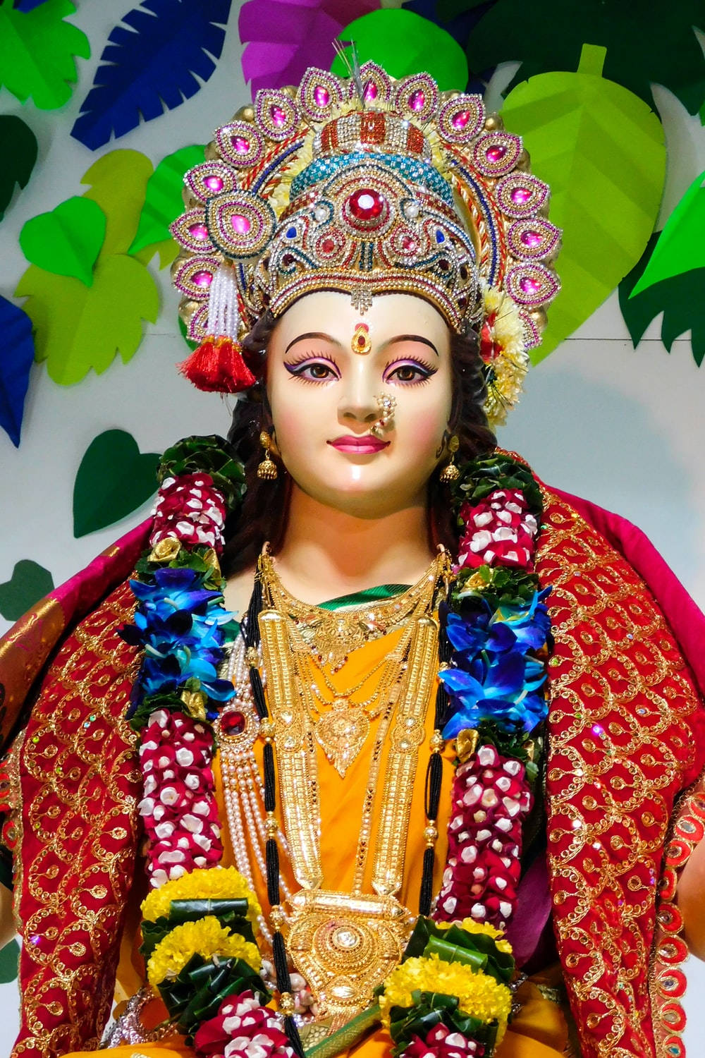 Vaishno Devi 桌布 壁紙 — 一位光芒四射的神祇，飾以精緻珠寶、色彩繽紛的花環和鑲有寶石的皇冠，背景是風格化的綠葉。