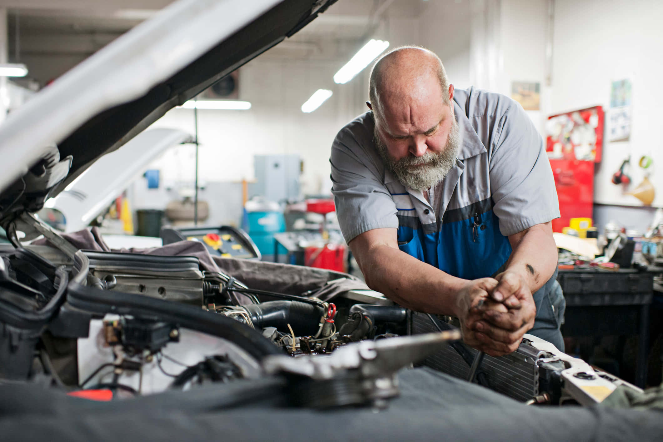 Car Repair 桌布 壁紙 — 一位光頭、留著灰色鬍鬚的技師在光線充足、工具齊全的車庫裡專注地修理汽車引擎蓋下的零件。
