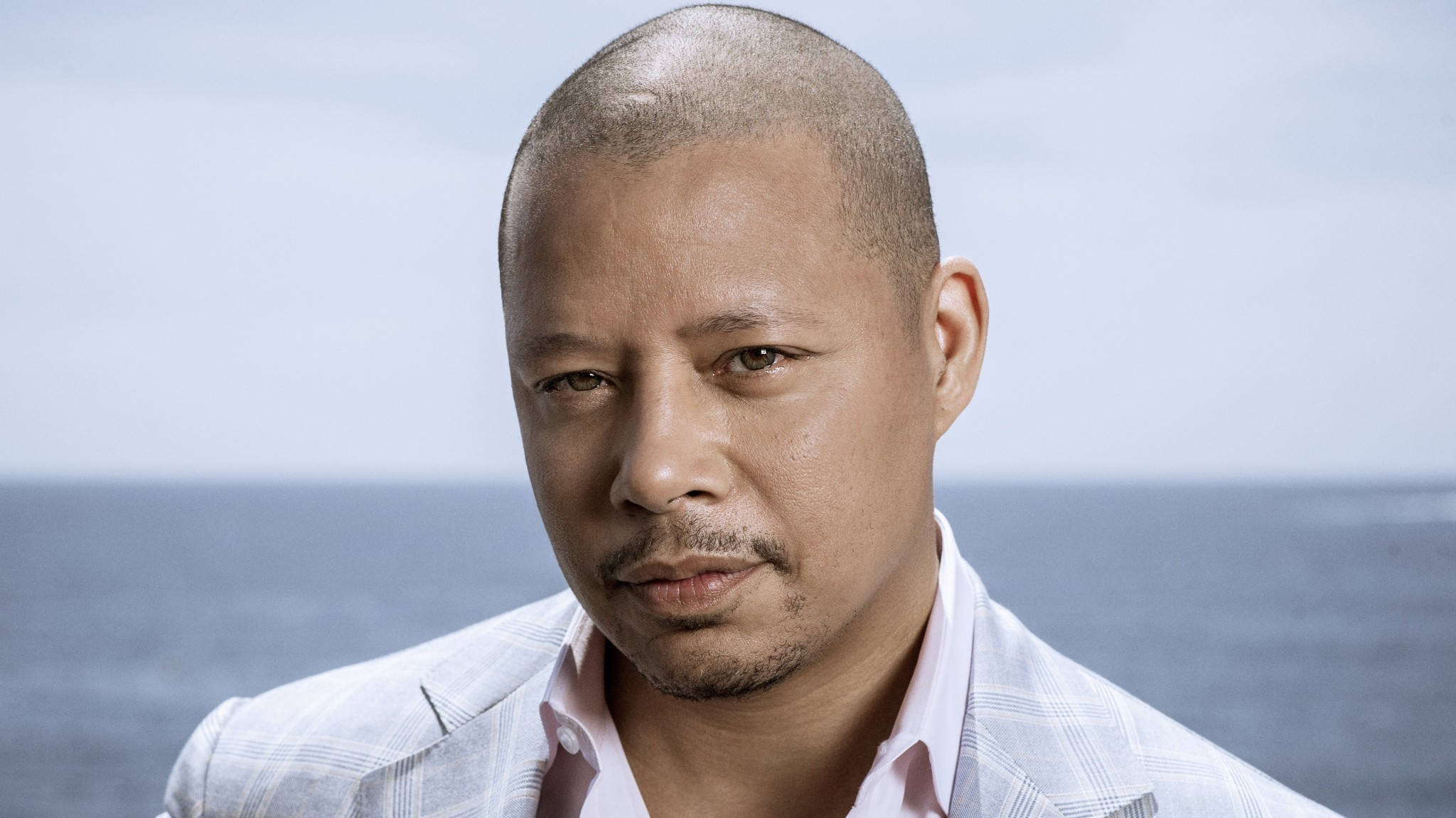 actor 桌布 壁紙 — 一位光頭、身穿淺色西裝的男子，在柔和日光下，以平靜的海洋為背景，若有所思地凝視著。
