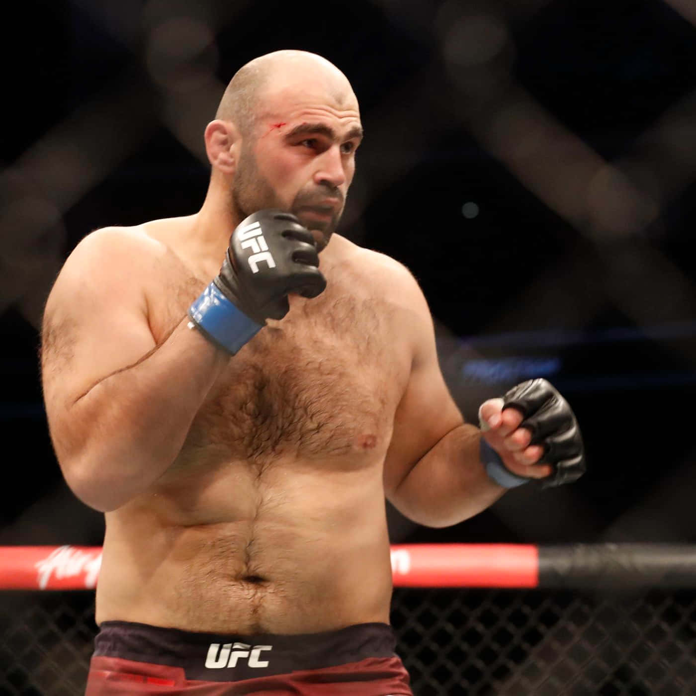 UFC 桌布 壁紙 — 一位光頭UFC拳手，眼睛流血，站在八角籠中，戴著黑色手套和紅色短褲。