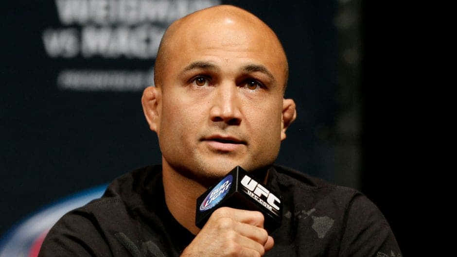 UFC 桌布 壁紙 — 一位光頭UFC拳手穿著黑色連帽衫，在模糊的宣傳背景的新聞發布會上對著麥克風講話。