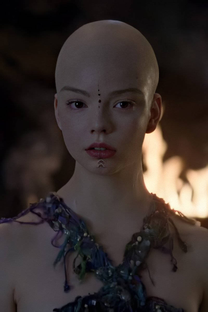 Anya 桌布 壁紙 — 一位光頭女性，深色眼睛，臉上有標記，穿著質感藍綠色上衣，背景溫暖發光。