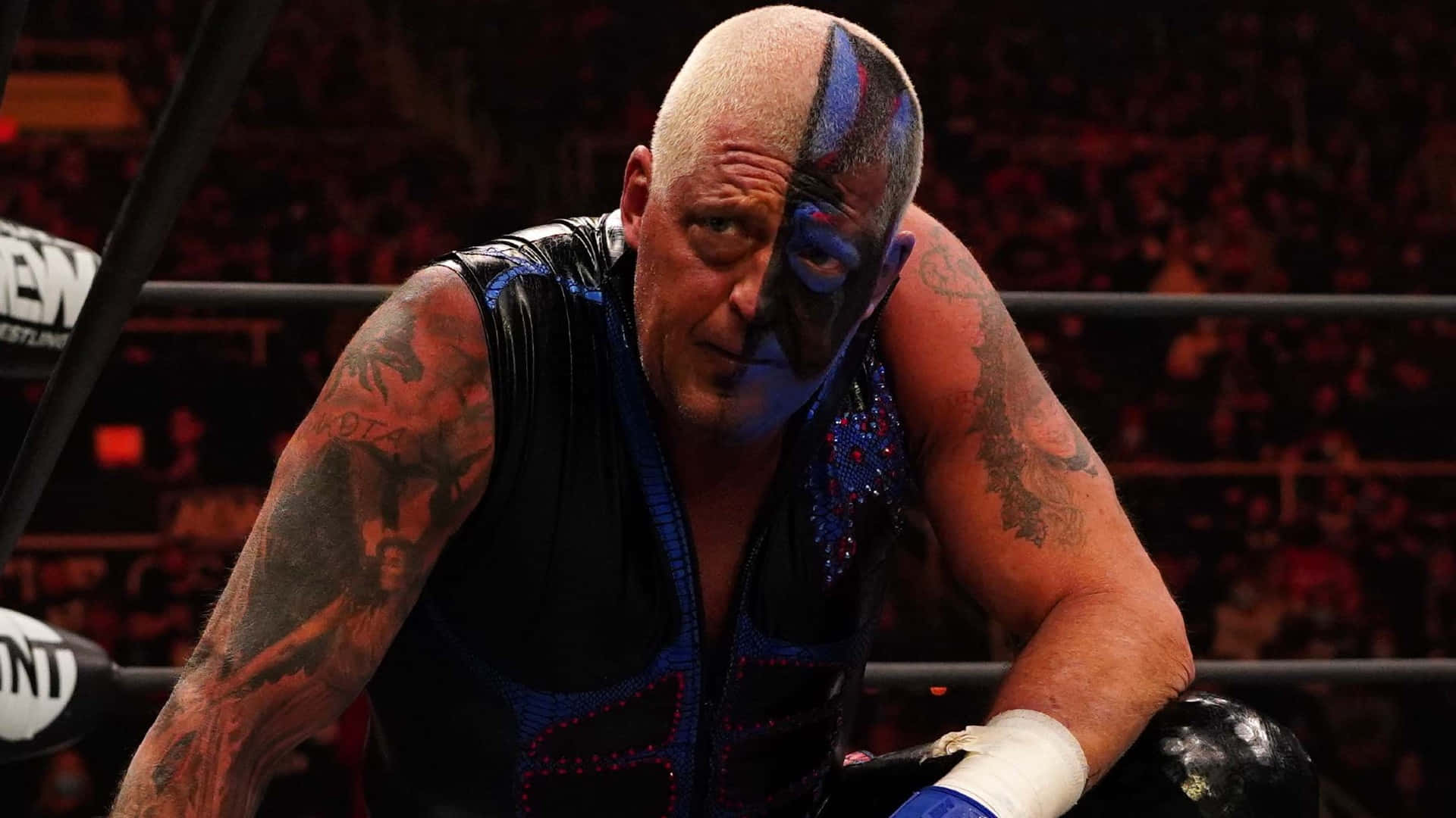 WWE 桌布 壁紙 — 一位光頭摔角手，臉上塗有藍色顏料和刺青，在明亮的場館燈光下靠在擂台上。