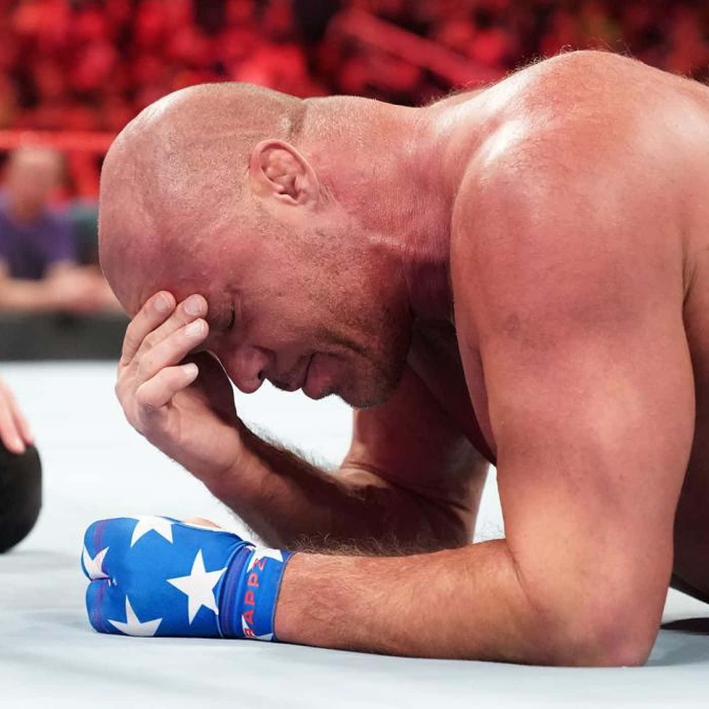 WWE 桌布 壁紙 — 一位光頭摔角手戴著藍色星星圖案手套，跪在墊子上，手托著頭，在紅色燈光的競技場中顯示失敗。
