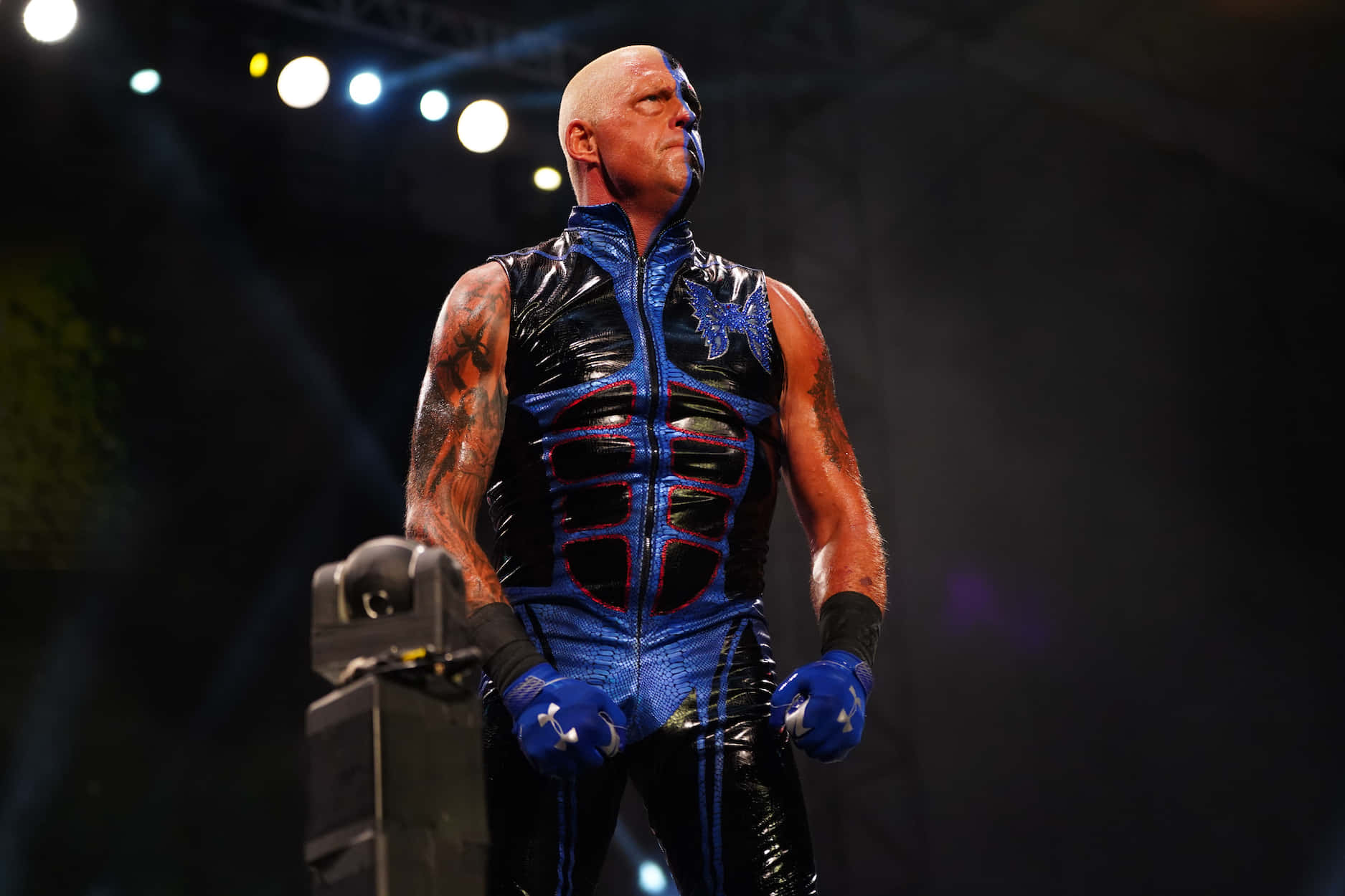 WWE 桌布 壁紙 — 一位光頭摔角手穿著閃亮的藍黑色服裝，在明亮的舞台燈光下自信地站立，手臂上的刺青清晰可見。