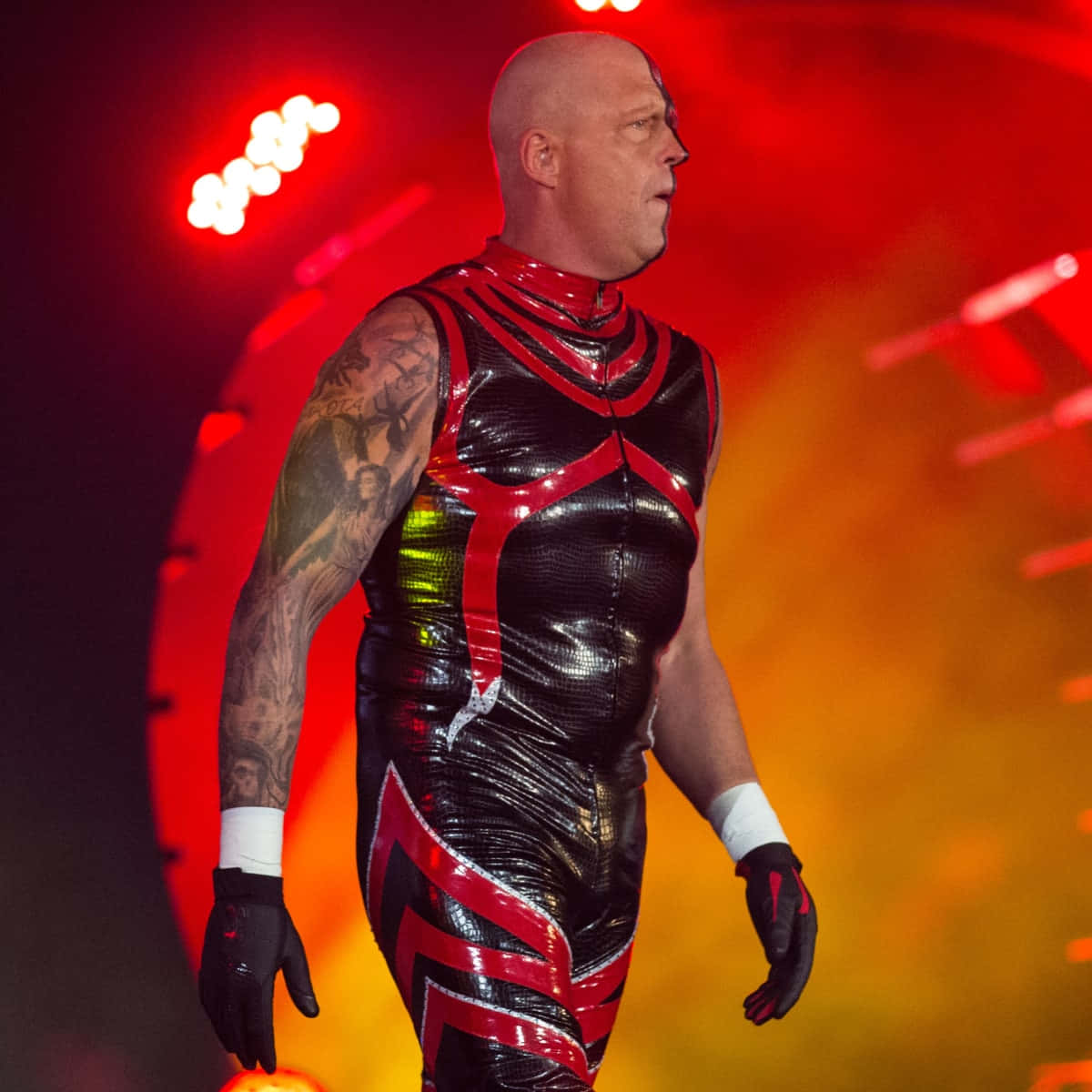 WWE 桌布 壁紙 — 一位光頭摔角手穿著閃亮的黑色和紅色緊身衣，身上有刺青，在紅色舞台燈光下行走。