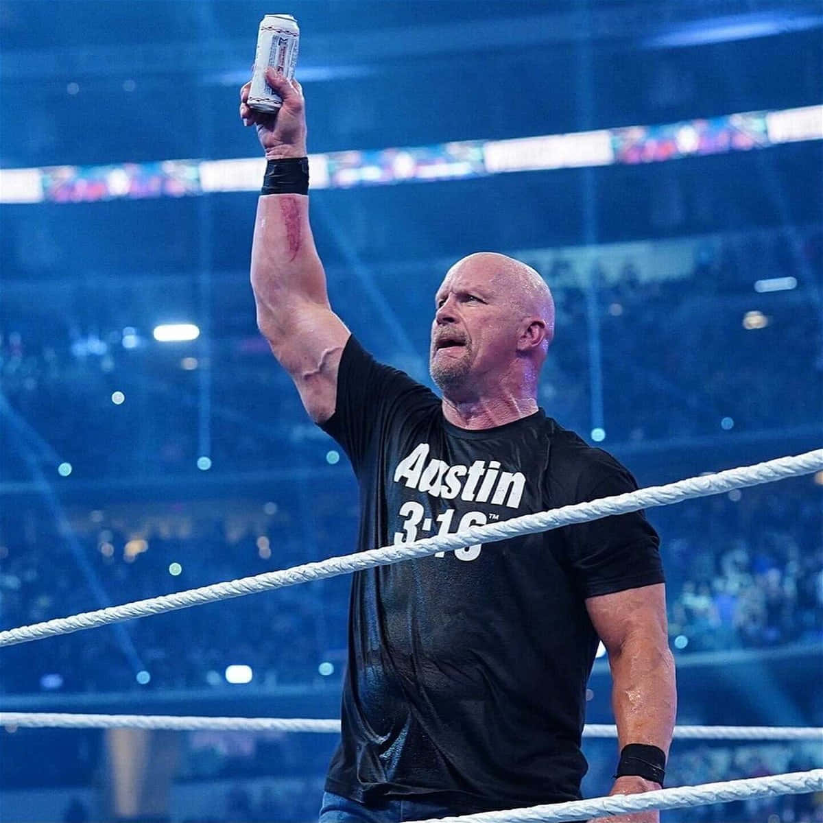 WWE 桌布 壁紙 — 一位光頭摔角手穿著黑色 'Austin 3:16' 襯衫，在燈光明亮的摔角場內舉起罐頭慶祝勝利。