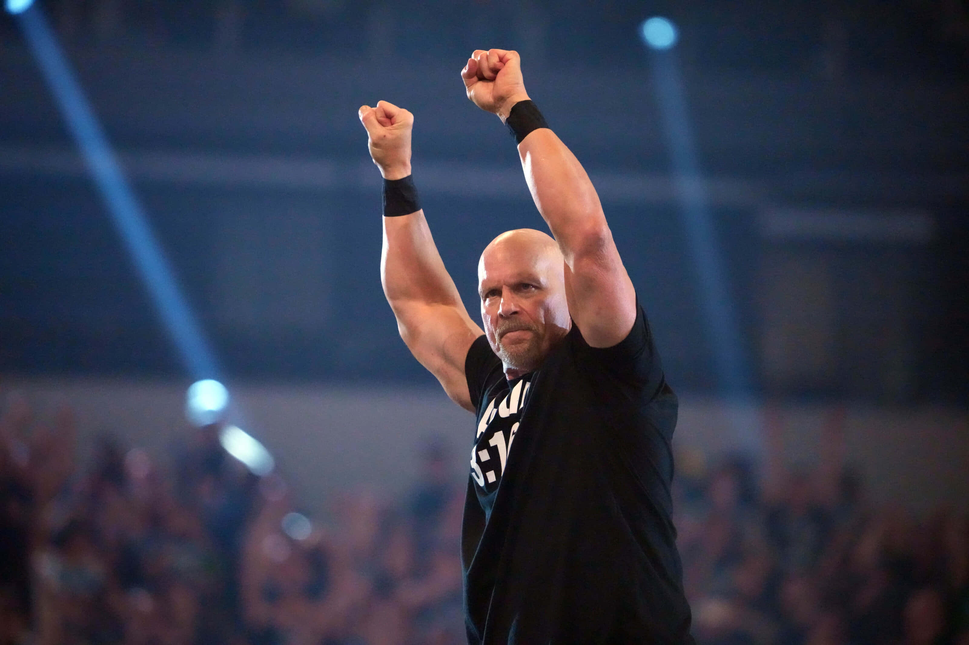 WWE 桌布 壁紙 — 一位光頭摔角手穿著黑色襯衫，在明亮的舞台燈光下高舉雙拳，身後是模糊的人群。