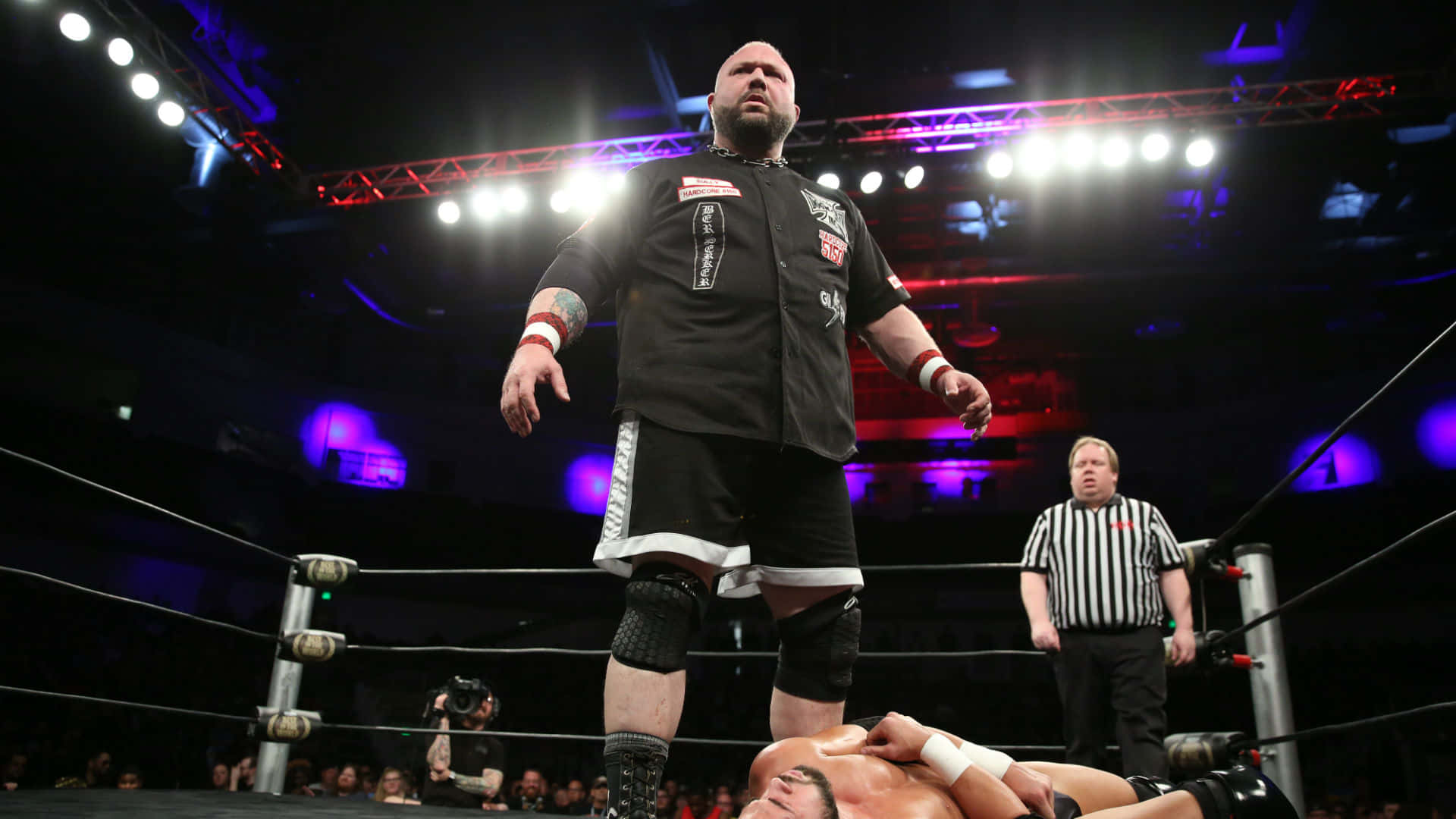 Wrestling 桌布 壁紙 — 一位光頭摔角手身穿黑色裝備，在燈光明亮的擂台上勝利地站在對手之上，裁判在旁觀看。