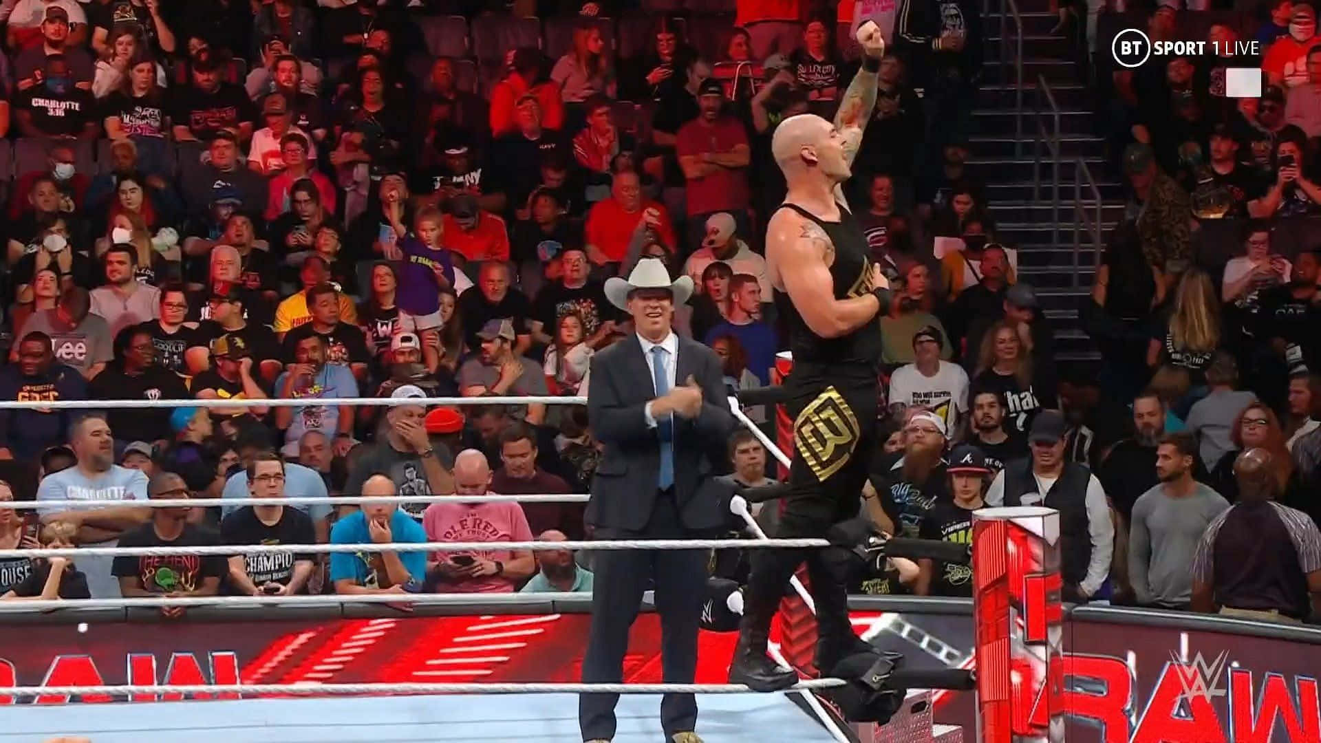 WWE 桌布 壁紙 — 一位光頭摔角手身穿黑色裝備在WWE擂台上勝利跳躍，一位穿西裝的播音員在咆哮的人群前鼓掌。