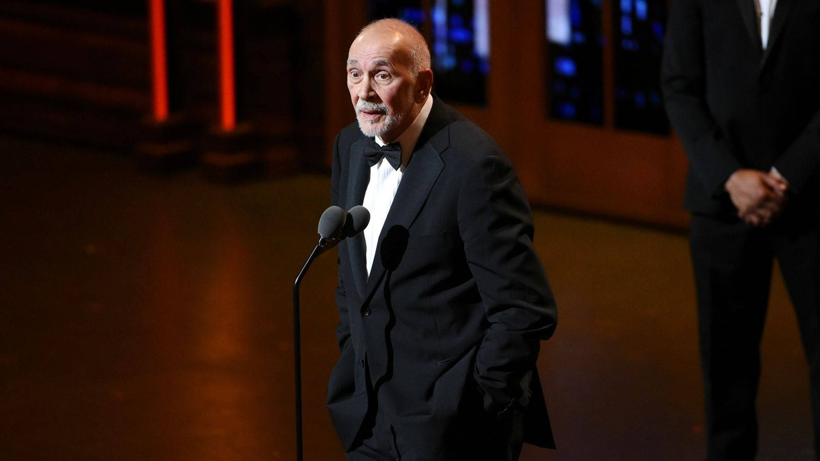 Celebrity 桌布 壁紙 — 一位光頭灰鬍鬚的男子身穿黑色燕尾服，站在昏暗的舞台上，舞台上有紅色和藍色的燈光，他正對著麥克風。