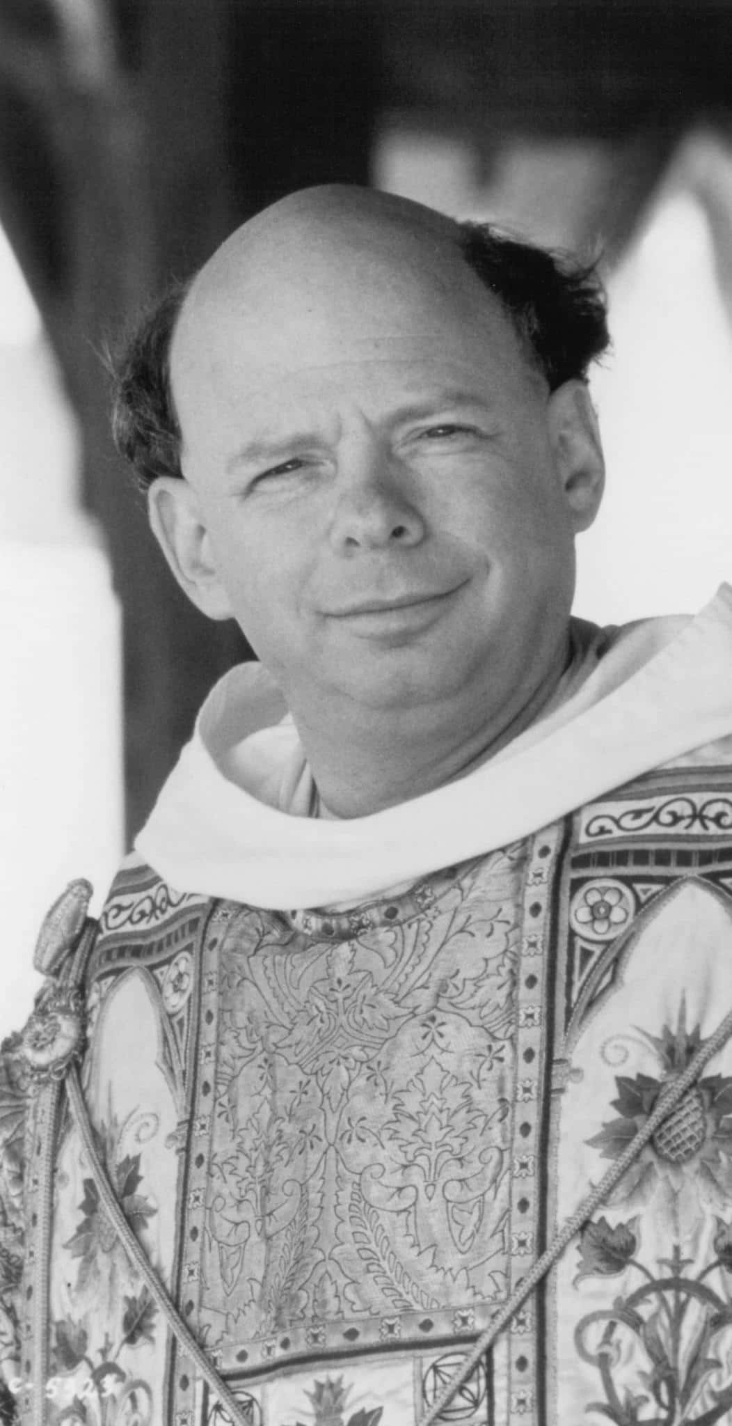 Wallace Shawn 桌布 壁紙 — 一位光頭男士的黑白肖像，面帶微笑，穿著帶有花卉和幾何刺繡的華麗儀式長袍。