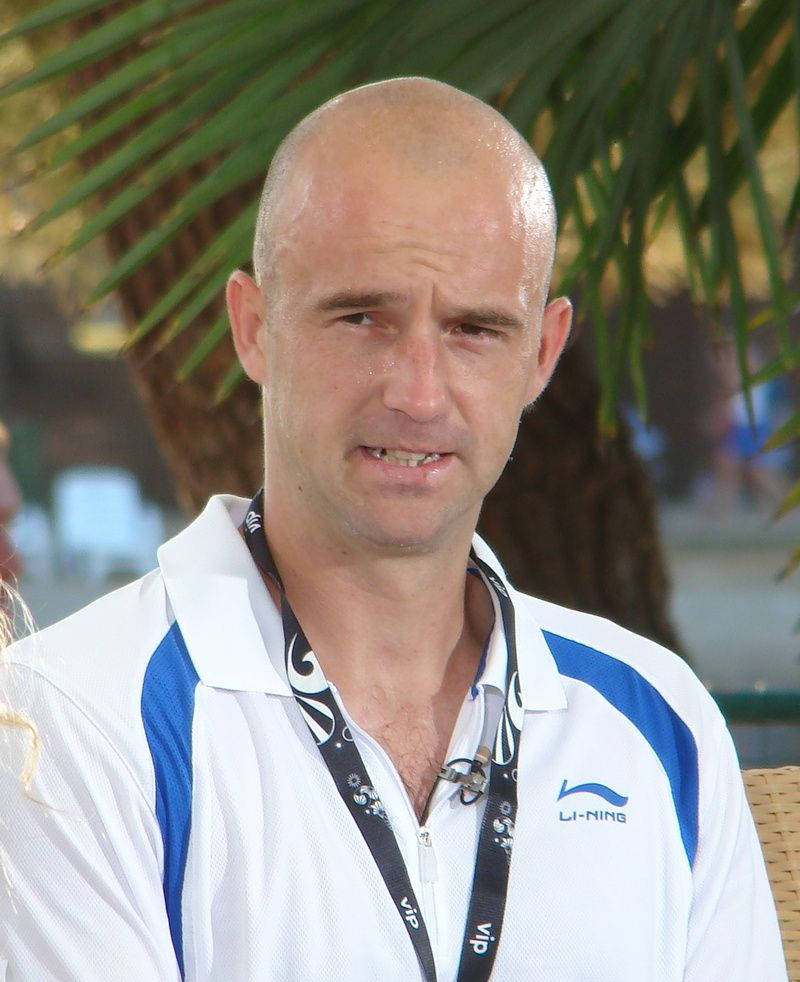 Tennis 桌布 壁紙 — 一位光頭男士穿著白色和藍色的Li-Ning Polo衫，掛著VIP證件帶，在棕櫚葉下顯得有點疲倦。