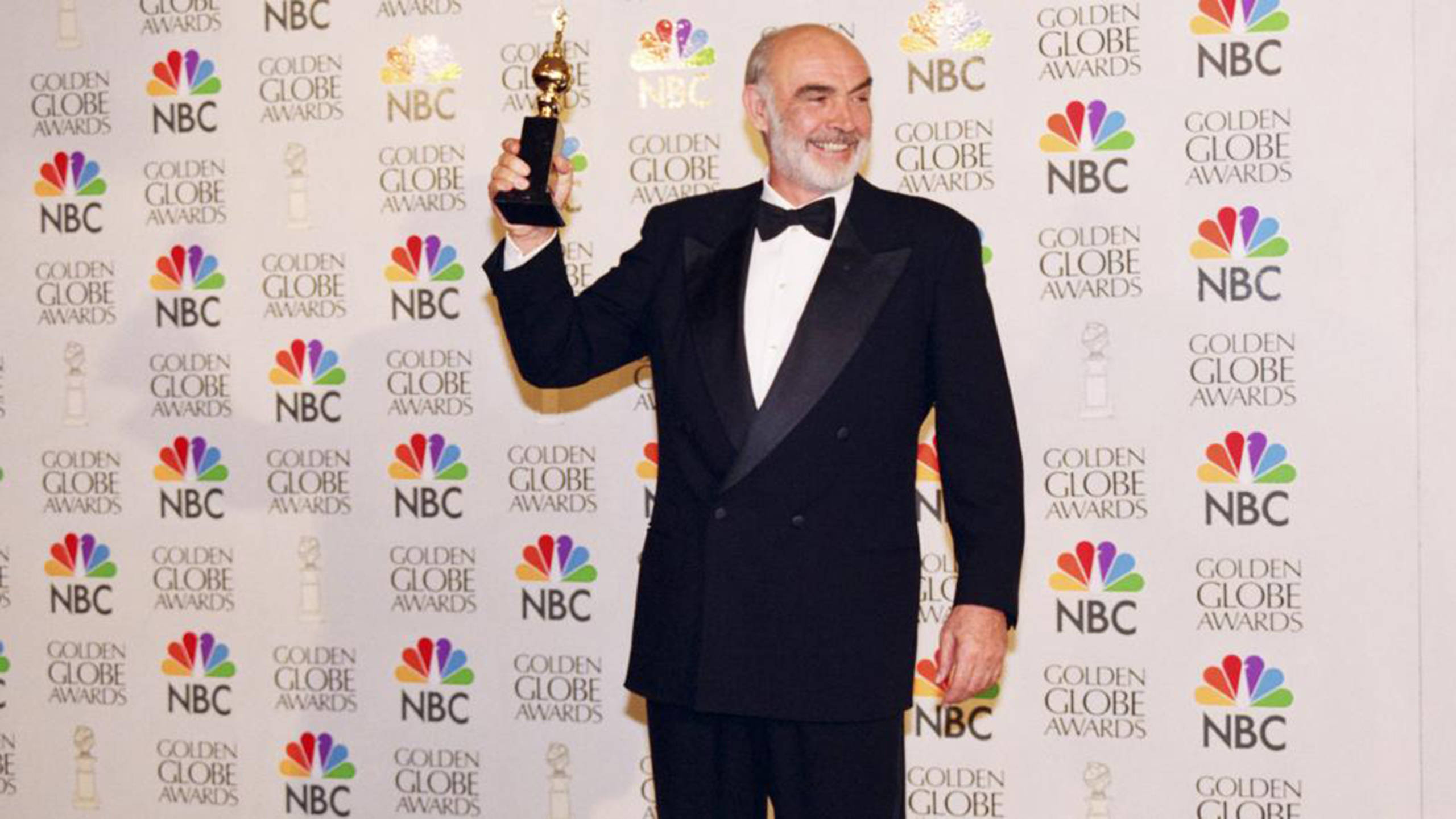 Sean Connery 桌布 壁紙 — 一位光頭男士穿著黑色燕尾服，在品牌活動背景前微笑著手持金色地球儀獎盃。