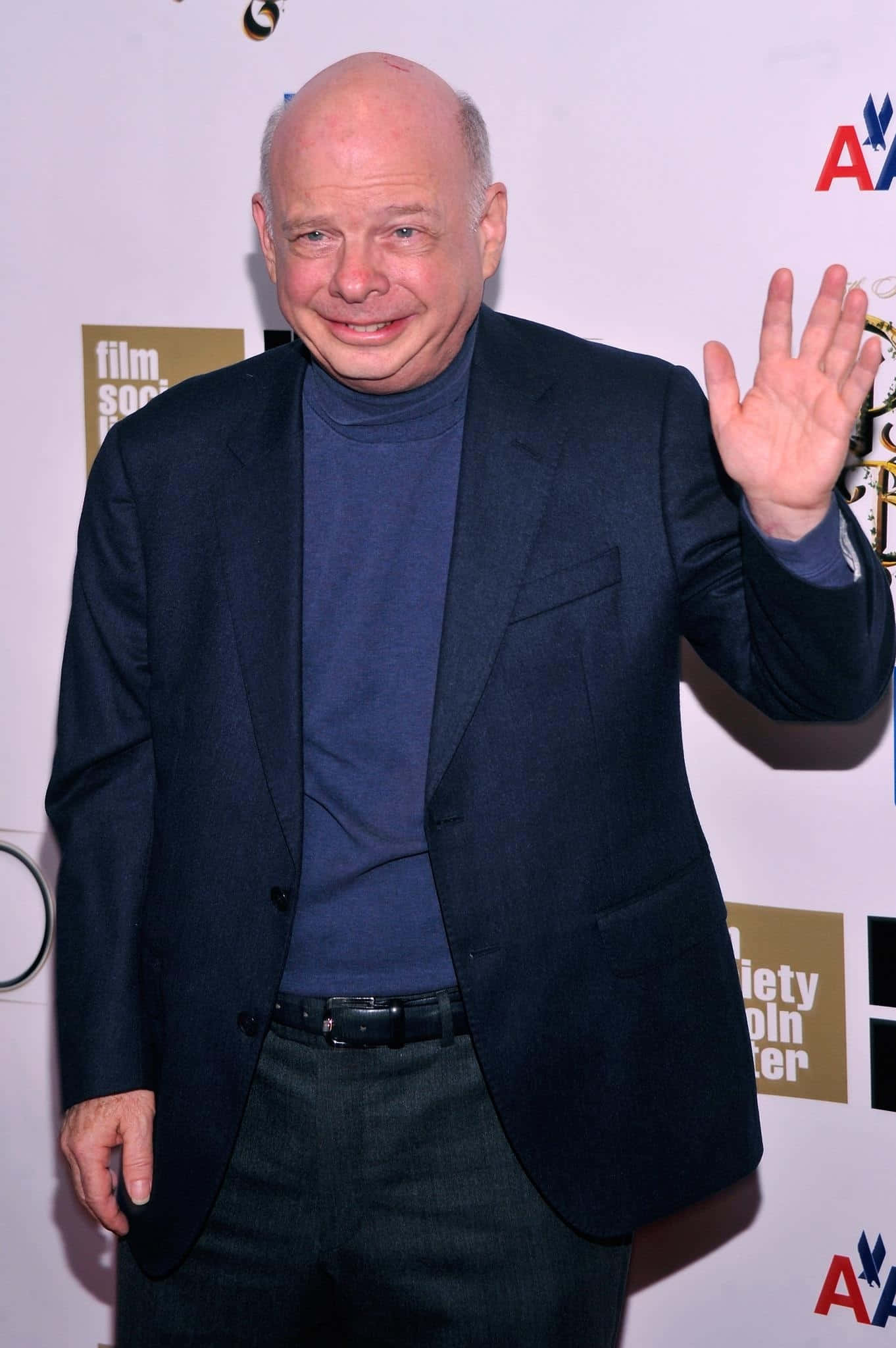 Wallace Shawn 桌布 壁紙 — 一位光頭男士身穿深色西裝和藍色高領毛衣，在背後有標誌的紅地毯活動上愉快地揮手。