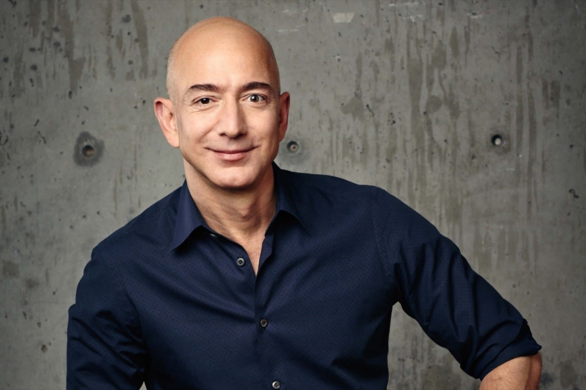 Jeff Bezos 桌布 壁紙 — 一位光頭男士身穿深藍色襯衫，在紋理灰色混凝土牆前自信地微笑。