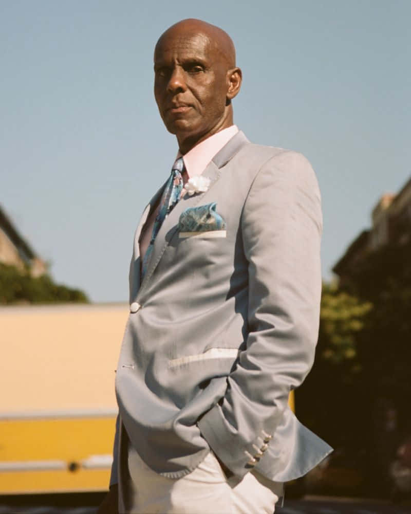 Man In Suit 桌布 壁紙 — 一位光頭男士身穿淺灰色西裝，搭配圖案領帶和口袋方巾，自信地站在晴朗藍天下的戶外。