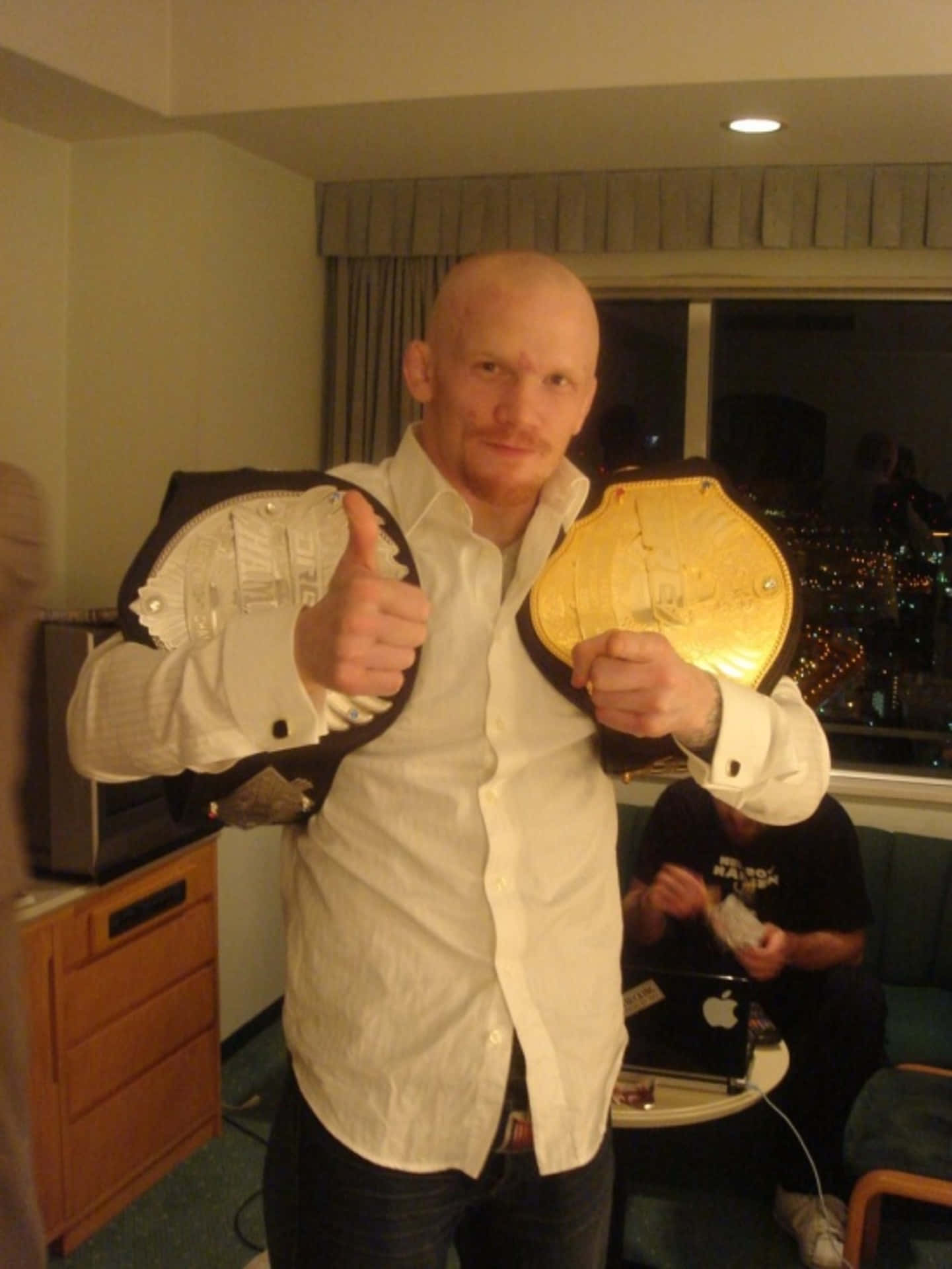MMA 桌布 壁紙 — 一位光頭男士身穿白襯衫，在夜晚的飯店房間內自豪地展示兩條冠軍腰帶並豎起大拇指。