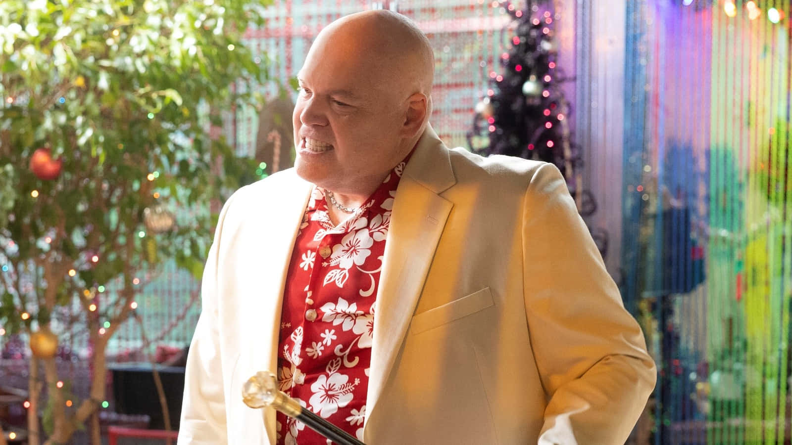 Vincent D'onofrio 桌布 壁紙 — 一位光頭男士身穿米色西裝和紅色花卉圖案襯衫，在節日色彩繽紛的環境中微笑著手持麥克風。