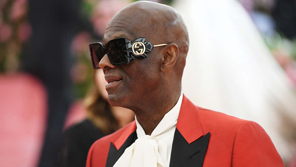 Gucci 桌布 壁紙 — 一位光頭男士身穿紅色燕尾服外套和白色領結，在一個華麗的活動中戴著超大的黑色Gucci太陽眼鏡。