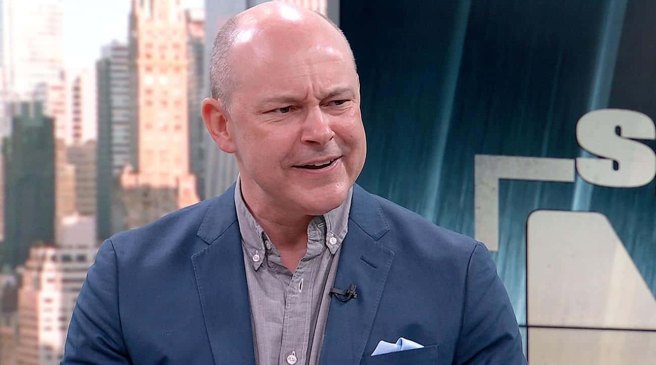 Rob Corddry 桌布 壁紙 — 一位光頭男士身穿藍色西裝外套，在模糊的城市天際線背景下於新聞佈景中微笑說話。