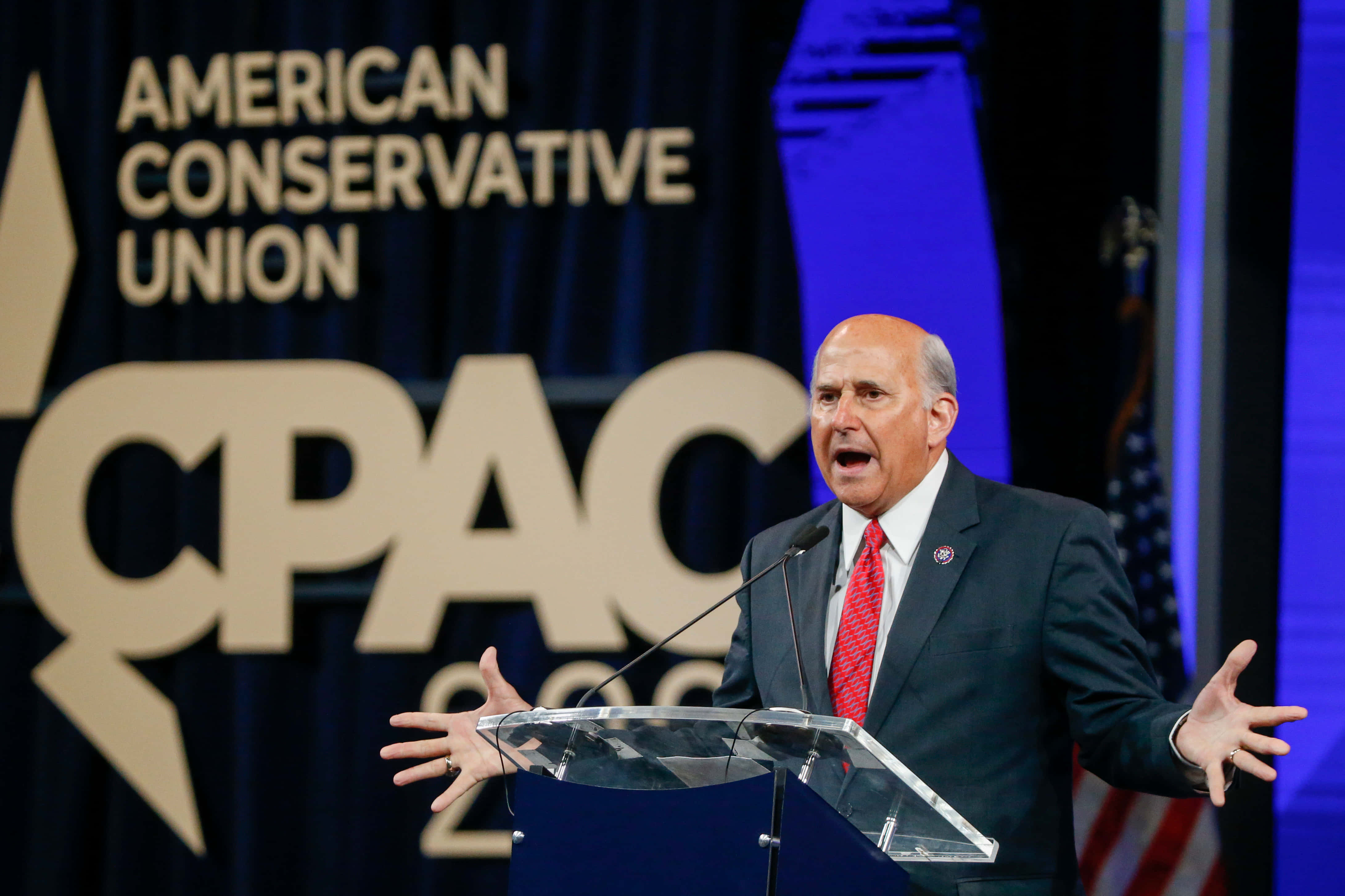 Man In Suit 桌布 壁紙 — 一位光頭男子身穿深色西裝和紅色領帶，在大型「CPAC」橫幅前的透明講台上熱情演講。
