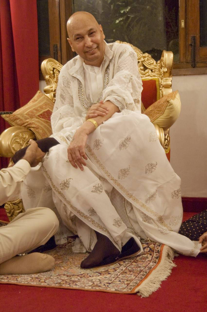 guru nanak dev ji 桌布 壁紙 — 一位光頭男子身穿白色刺繡服裝，坐在金色寶座椅上，身後有紅色窗簾，露出溫暖的笑容。